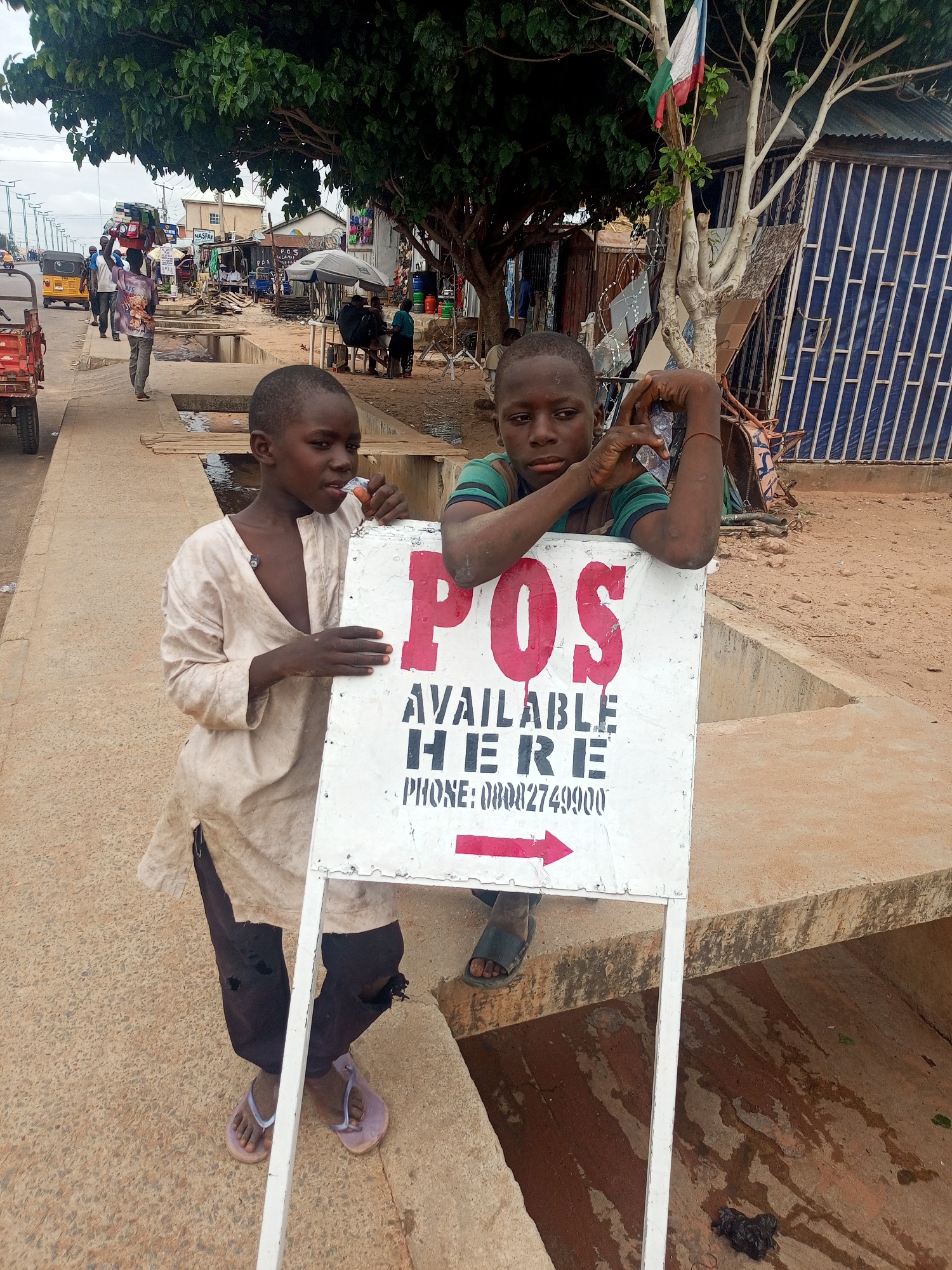 The location is an urban street scene in a city in Unknown, Unknown.
The image captures two young, dark-skinned boys in the foreground, positioned in front of a white A-frame sign. The boy on the left, wearing a loose-fitting, light-colored long-sleeved tunic over dark pants and light purple flip-flops, holds the sign with his right hand and appears to be eating a small, orange-colored item with his left hand, looking slightly to the left of the camera. The boy on the right, clad in a green and dark-striped short-sleeved shirt and dark flip-flops, leans on the top edge of the sign with both forearms, hands clasped, and rests his chin, looking directly at the camera with a serious expression.
The sign prominently displays "POS" in large red letters, followed by "AVAILABLE HERE" in black, and "PHONE: 08082749900" with a red arrow pointing right. The sign itself appears weathered with some paint drips.
Behind the boys and the sign, an active street scene unfolds. To the left, a paved road shows light vehicle traffic, including a yellow auto-rickshaw. Pedestrians walk on a concrete sidewalk; one man is visible carrying a stack of boxes or containers on his head, moving away from the camera. The sidewalk runs parallel to a concrete drainage ditch.
To the right and in the background, a cluster of makeshift market stalls and structures made of corrugated metal and other materials are visible. Several people are gathered under a gray market umbrella. A large tree with dense green foliage provides shade on the left side, while a second tree with sparser branches on the right features a small flag with horizontal stripes of green, white, and red tied to one of its branches. Other details include signs for "NASFAY" and "P.O.S SALA" (partially visible), and red gas cylinders. The ground is a mix of concrete pavement and packed dirt/sand, with a small pile of dark refuse near the base of the sign on the right. The sky overhead is overcast.