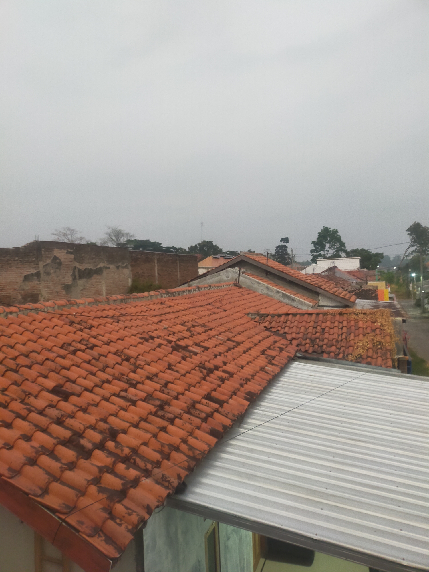 The image shows a view of rooftops in Tegalandong, Indonesia, under an overcast sky. The foreground is dominated by numerous orange clay roof tiles arranged in overlapping rows. To the left, a weathered brick wall is visible, with some sections showing signs of disrepair and exposed mortar. Beyond the immediate rooftops, a variety of other roofs can be seen, some with similar orange tiles and others with different materials, including what appears to be a corrugated metal roof in the lower right portion of the image. In the background, there are trees and some distant buildings, suggesting a residential or semi-urban environment. The overall impression is that of a residential area on a cloudy day. There are no people visible in the image.