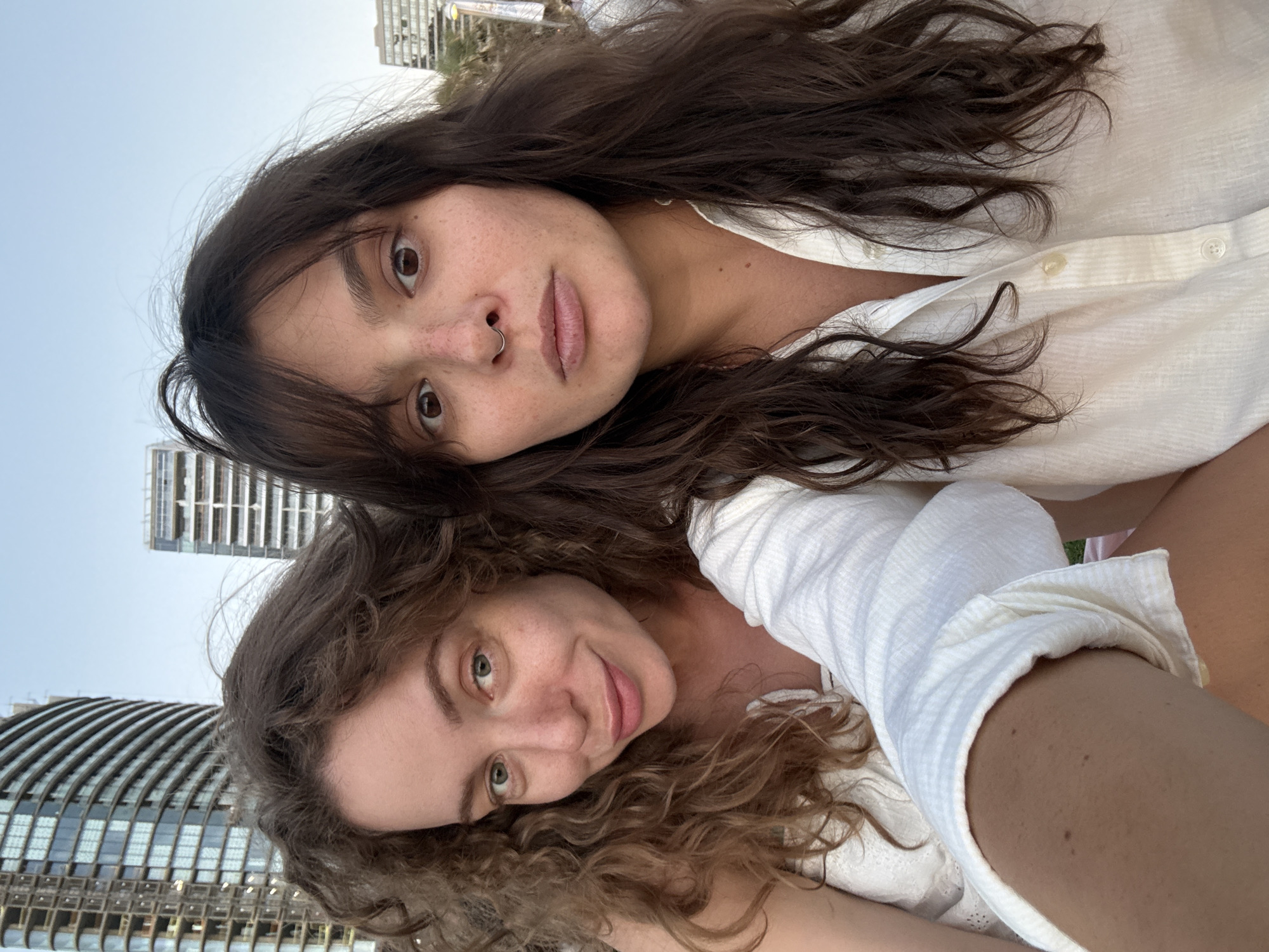 Two women are posing for a selfie outdoors. The woman on the left has curly brown hair and light green eyes, and she is smiling. The woman on the right has dark brown hair and brown eyes, and she is looking at the camera with a neutral expression. Both women are wearing white tops. In the background, there are several modern buildings under construction, suggesting an urban environment. The overall scene is a candid shot taken in Tel-Aviv, Israel, likely in an area experiencing development.