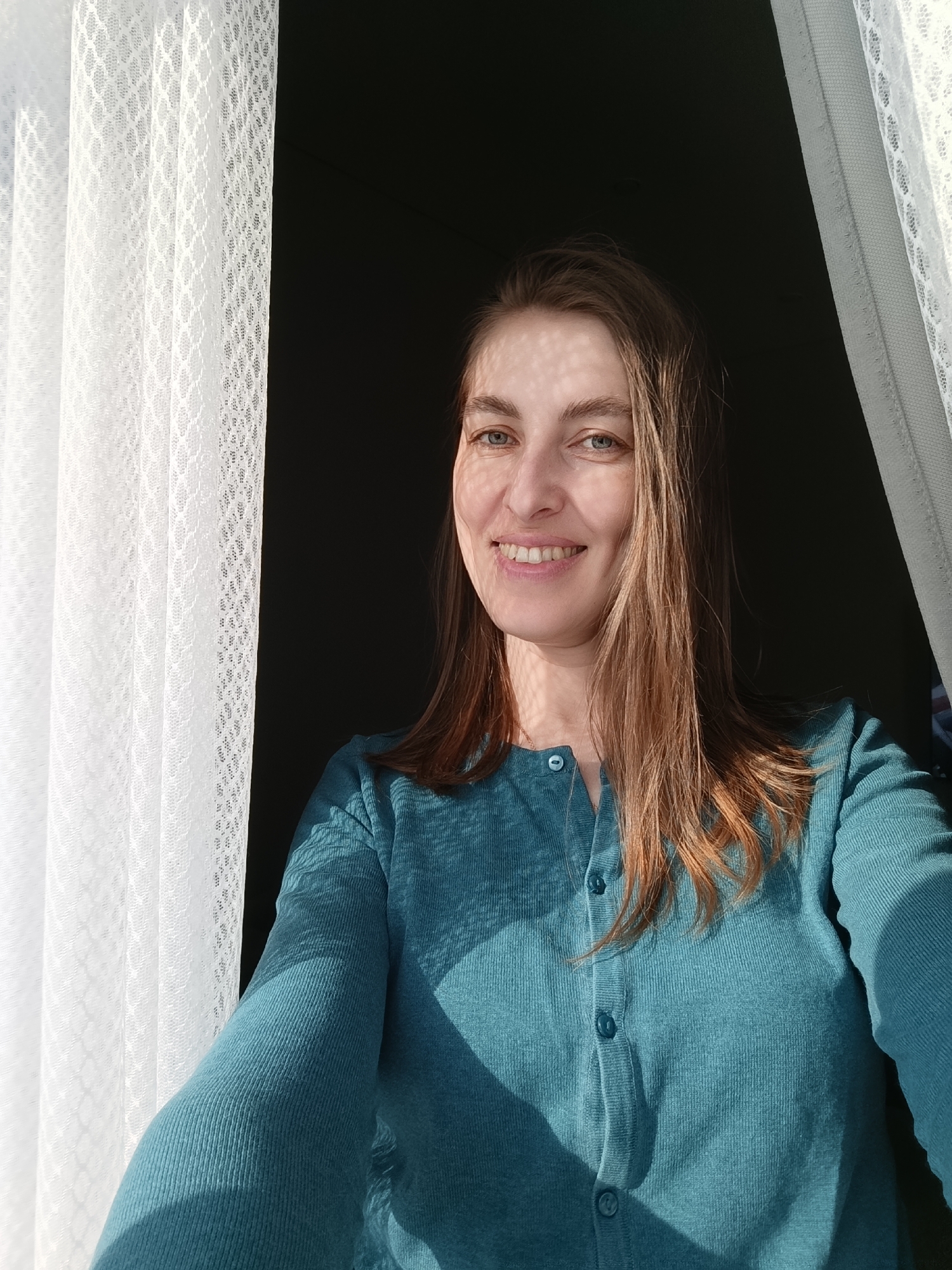 A woman with long brown hair, blue eyes, and a bright smile poses for a selfie in front of a window. She is wearing a turquoise button-down sweater. The background behind her is dark, suggesting an interior space. Light filters through the white lace curtain to her left, casting patterned shadows on her face and clothing. The location is Chaikovsky, Russia.