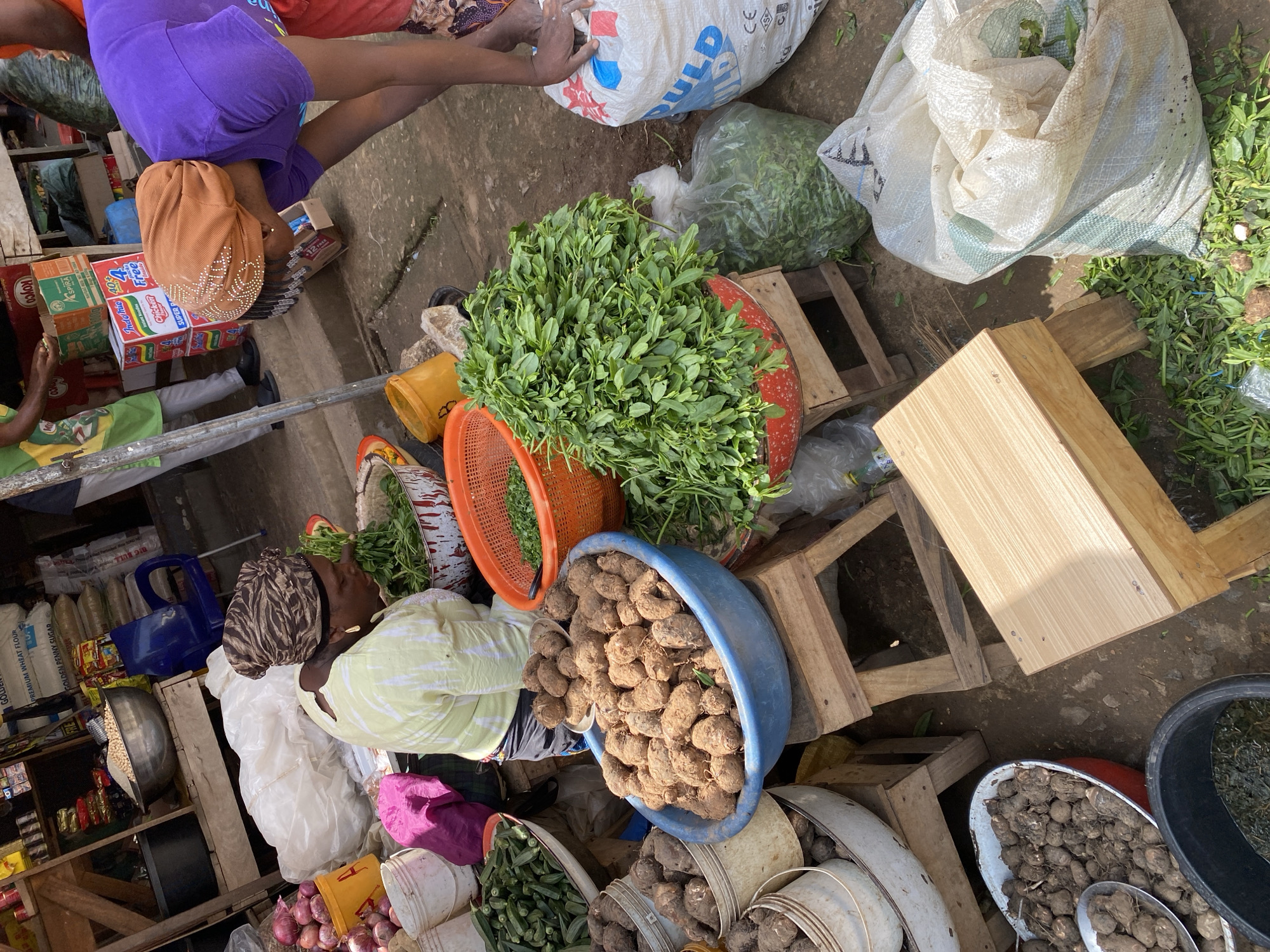An outdoor market scene in Jos, Nigeria, shows several individuals engaged in commerce. A woman wearing a yellow shirt and a patterned headscarf is sorting green leafy vegetables in a red and white patterned bowl. To her right, another person in a purple top and orange headscarf is reaching towards a large white sack, possibly a customer or vendor. Several stalls display a variety of fresh produce, including tubers in blue and red bowls, okra in a white bowl, and bundles of green leafy vegetables in red baskets. Bags of produce and other goods are visible, along with wooden crates and makeshift shelving. The ground is dirt, with some debris and scattered produce. The overall atmosphere is that of a bustling marketplace.