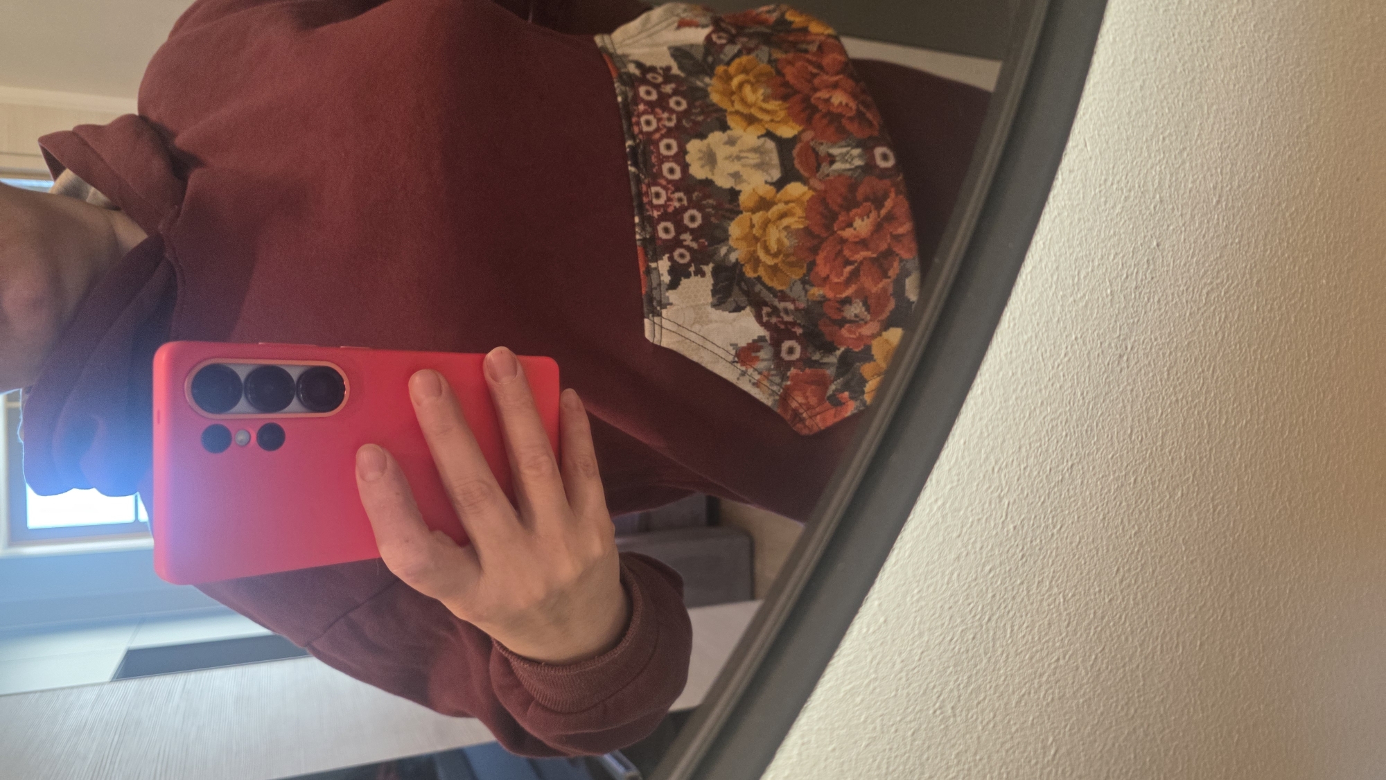 A person takes a selfie in a mirror. The person is wearing a maroon-colored hoodie with a floral pocket. The phone in the person's hand is in a pink case. The location is not identifiable as Saint Petersburg, Russia, or any specific city.