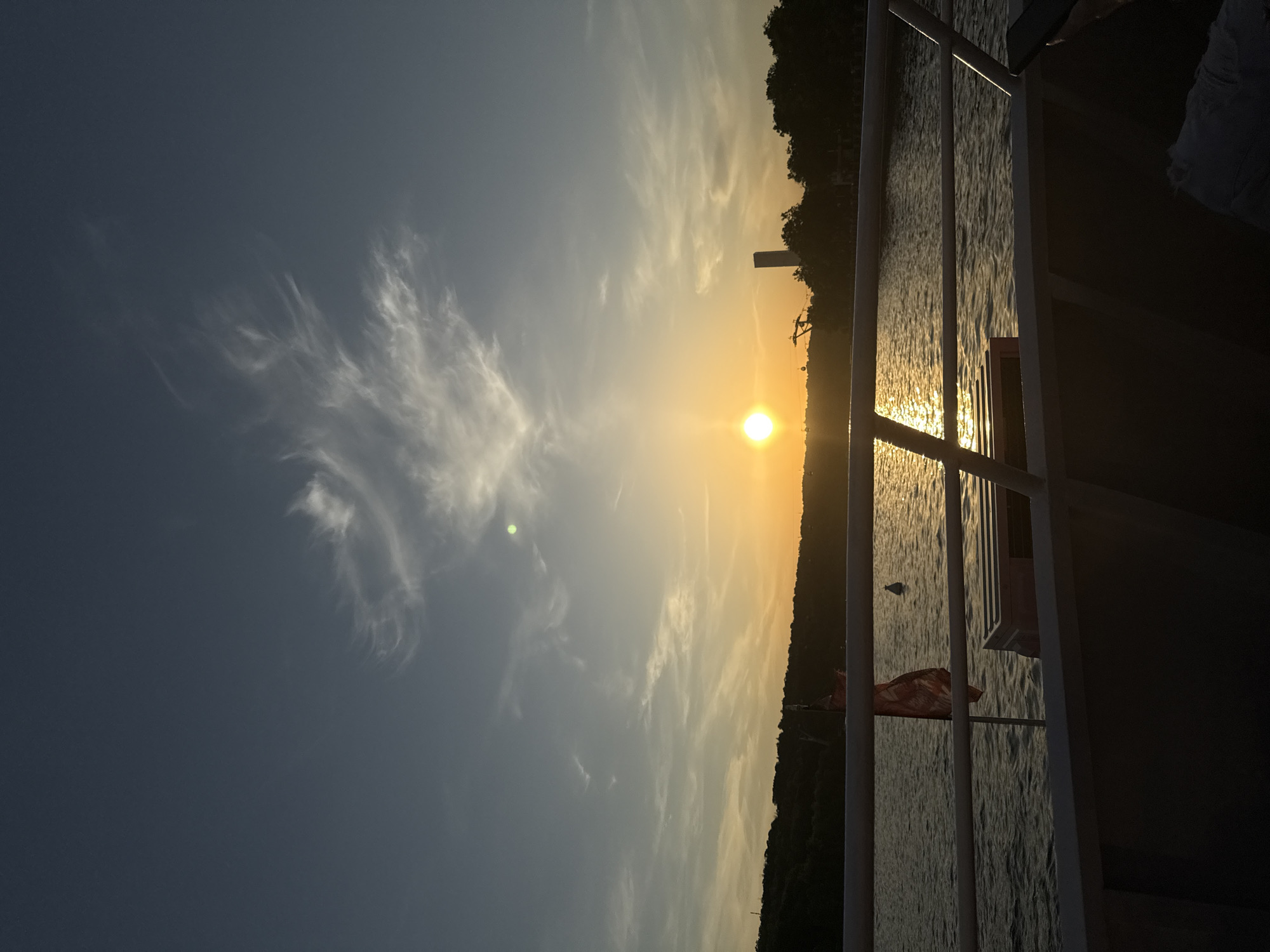 The image depicts a sunset over water, viewed from what appears to be a boat or ship, with railing visible in the foreground. The sun is positioned on the horizon, casting a bright glow and reflections on the water. The sky shows gradient hues of blue and orange, with some cloud formations. The opposite bank of the water body is visible as a dark silhouette of trees and vegetation, punctuated by the silhouette of structures. The image is underexposed. The city in the location cannot be known without more details.