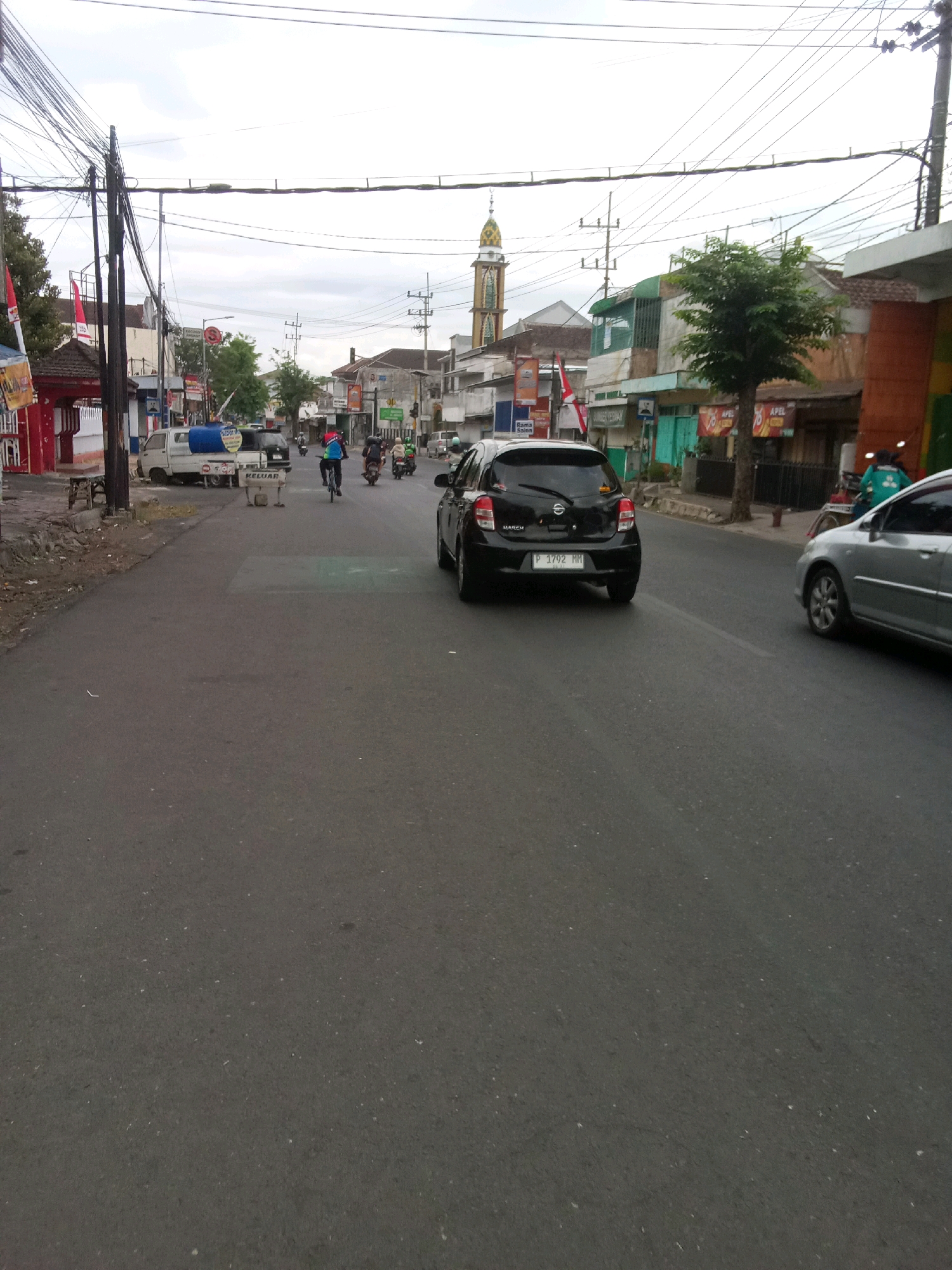 The image depicts a street scene in Jember, Indonesia. The road is asphalt, and there are buildings and vegetation lining the sides. Several vehicles are present, including cars, motorcycles, and a white utility truck. People are seen riding motorcycles and a bicycle. Overhead, power lines crisscross the sky, and a minaret of a mosque is visible in the distance. The weather appears overcast. Signs in Indonesian are visible on some of the buildings, and one sign on the utility truck says "KELUAR," which means "EXIT" or "OUT."