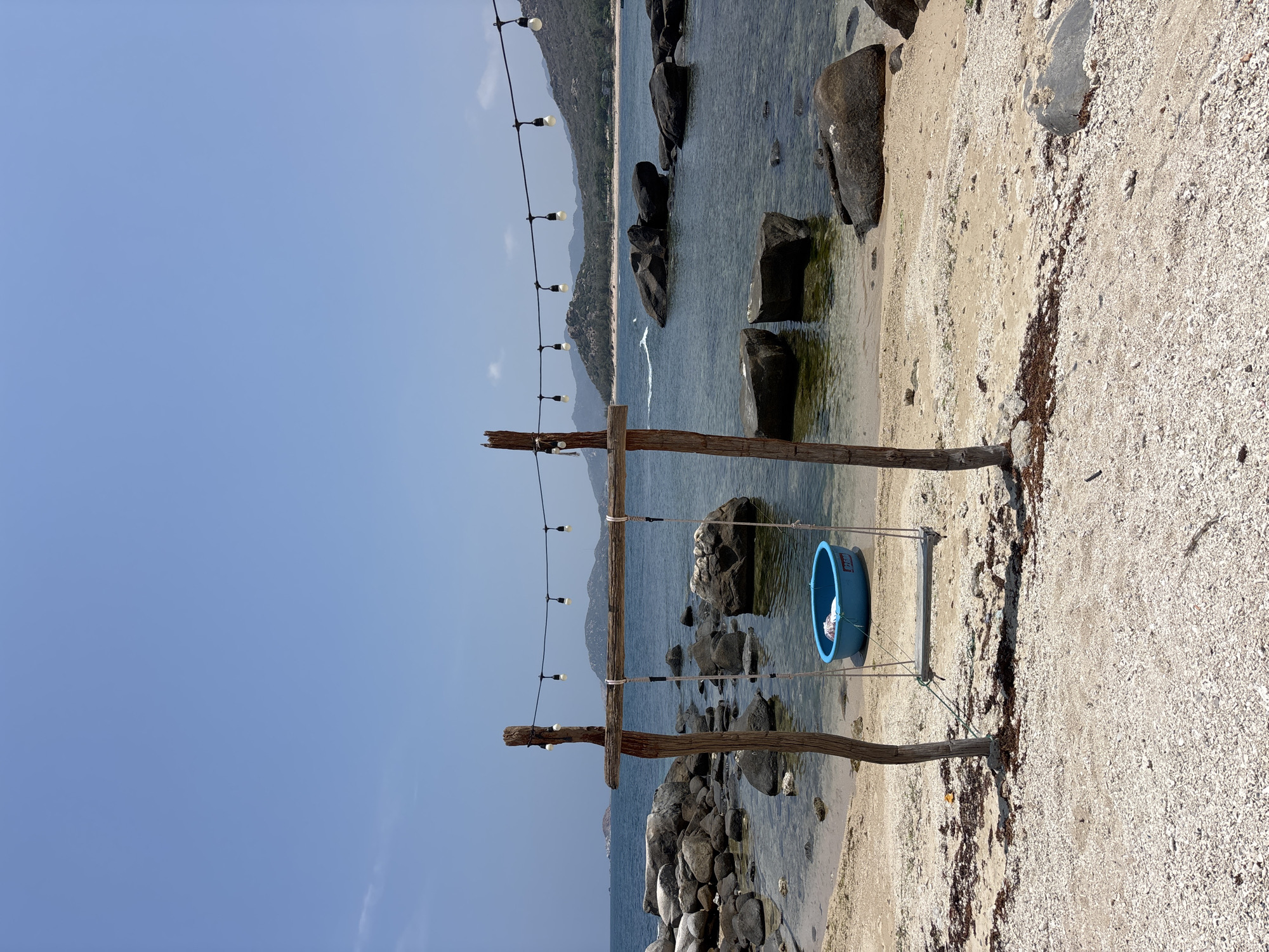 A landscape shot depicts a crude swing, featuring a large blue plastic tub suspended by ropes from a makeshift wooden frame. The swing is positioned on a beach, where the ground is a mix of sand and small rocks, with scattered seaweed. Behind the swing, the frame is illuminated by a string of lights strung along the wooden beams. The scene transitions to a body of water dotted with large rocks emerging from the surface. In the distance, the vague silhouette of a forested mountain range is visible, set against a clear, blue sky. This landscape is located in Vinh Hai, Vietnam.