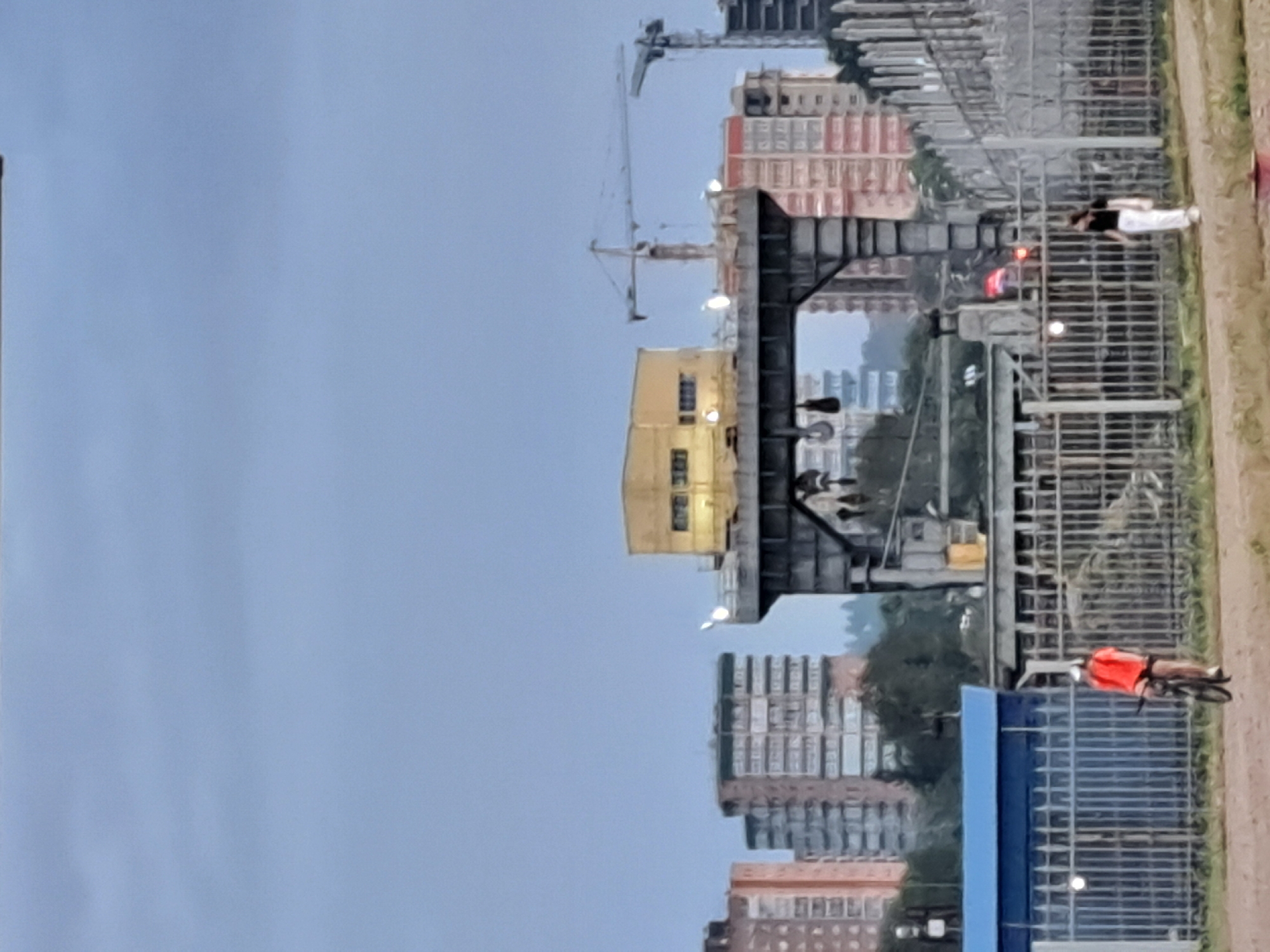 This image depicts an outdoor scene with a city skyline in the background, featuring apartment buildings and construction cranes. In the foreground, a dirt path runs alongside a metal fence. A person on a bicycle, wearing an orange shirt and black shorts, is positioned on the path. Further along the path, another person in a black top and white pants is walking. The overall lighting suggests it is either early morning or late evening. The scene appears to be within the city of Irkutsk, Russia, potentially near a construction site or industrial area given the presence of cranes and the bridge-like structure in the mid-ground.