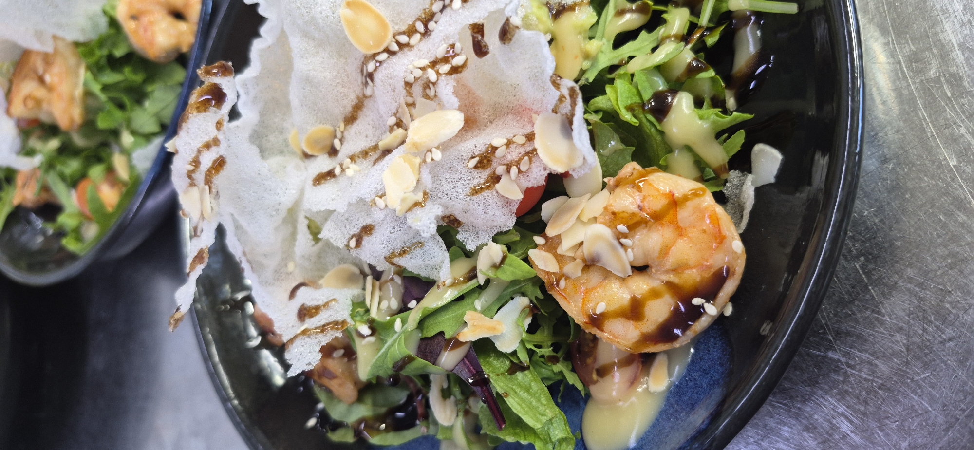 A close-up, eye-level shot shows a gourmet salad served in a dark blue bowl, situated on a scratched metal surface. The salad features a prominent, glistening shrimp coated in a dark sauce and sprinkled with sliced almonds and sesame seeds. It also contains mixed greens, a white crispy cracker-like garnish drizzled with a similar dark sauce, and dollops of light-colored dressing. The background shows another bowl with a similar salad, out of focus. The lighting is bright and direct, creating reflections and highlights on the food and bowl. The scene appears to be an indoor setting, possibly a restaurant kitchen or dining area. There is no visible indication of the city of Saint Petersburg, Russia.