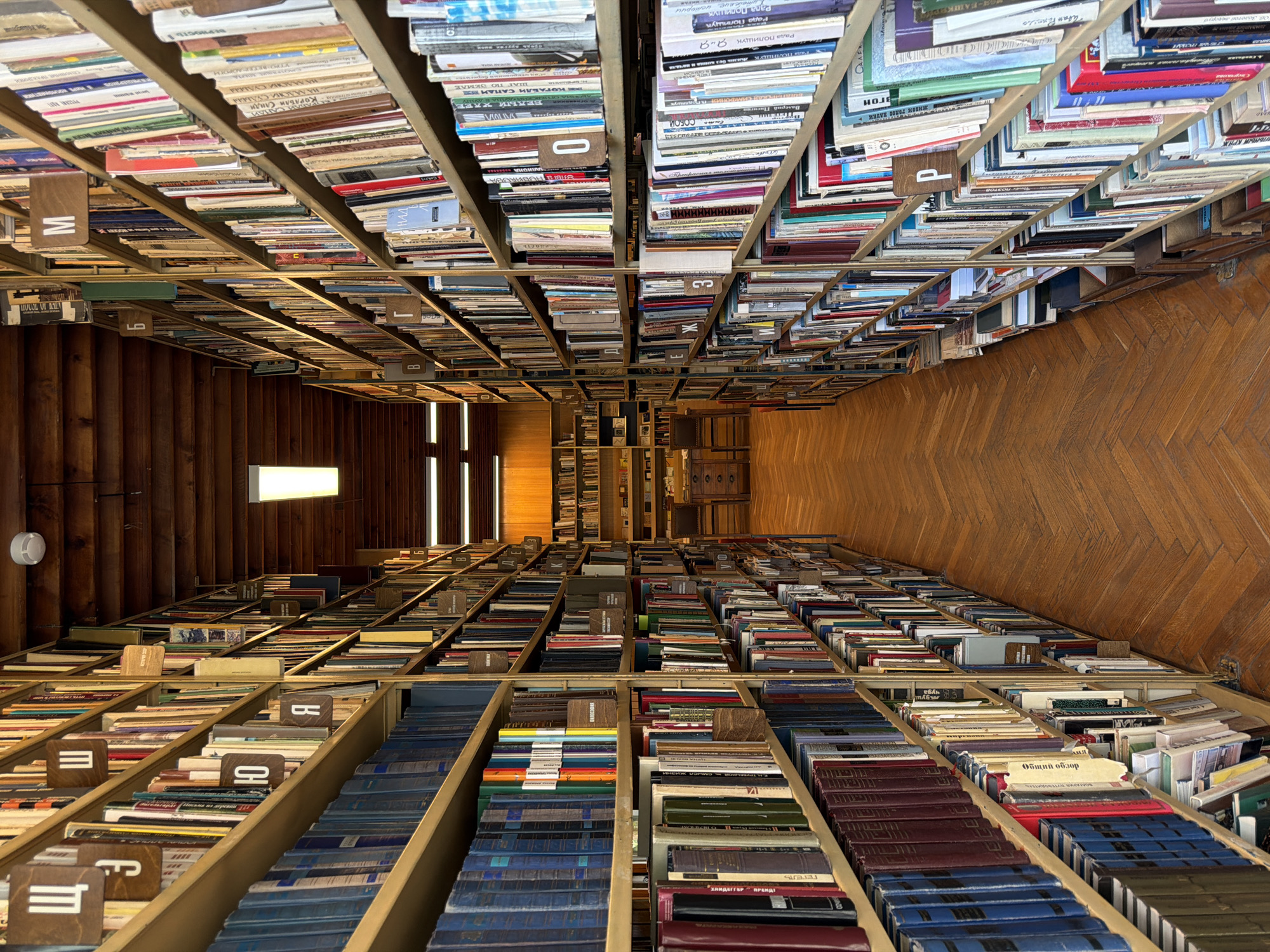 Here is a detailed description of the image:
The image showcases an interior view of a library or book repository, likely taken from a raised perspective looking downwards. The space is filled with bookshelves laden with books, creating an immersive and dense arrangement. The books are of varying sizes, colors, and ages, suggesting a diverse collection.
Lettered shelf markers are visible throughout the photo, aiding in the organization of the books.
The floor appears to be wooden, laid out in a parquet or herringbone pattern. In the background, there is a set of steps leading upwards, presumably to other levels of the library. There is a light fixture emanating soft light on one part of the staircase.
The arrangement and scale of the bookshelves create a feeling of depth and perspective within the image.
I can infer that the location is in the city of Vnukovo, Russia.