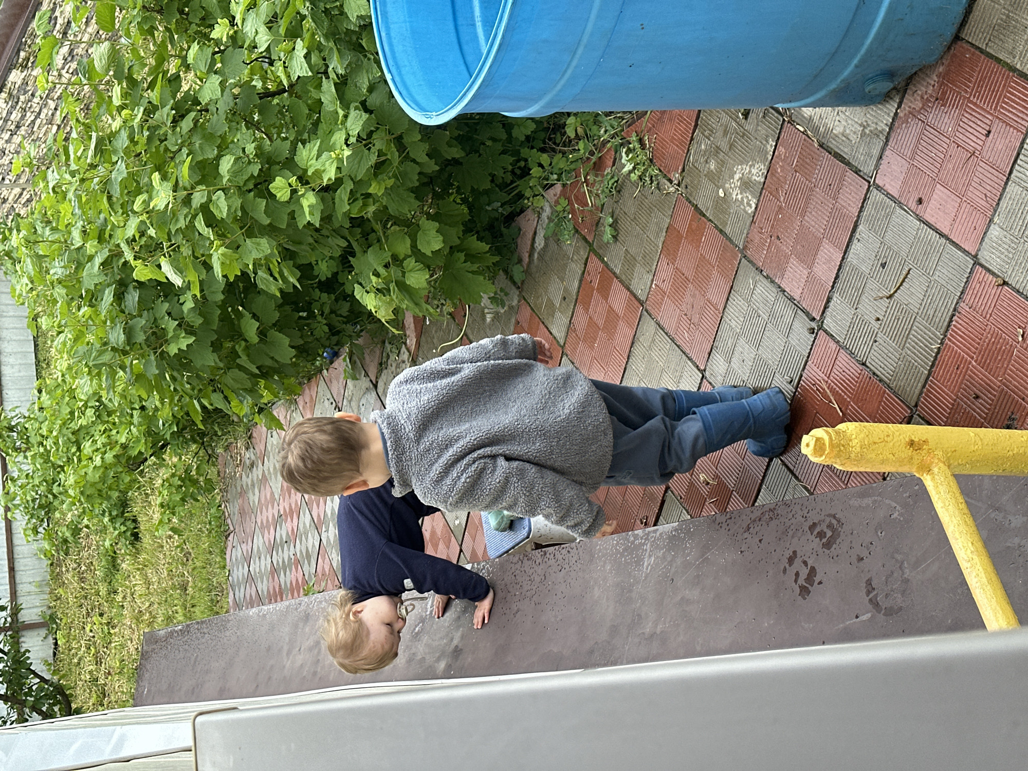 Two children are outside on a gray surface with dark brown paw prints. The child in the foreground is younger and wearing a dark blue shirt and pants. The older child is wearing a gray fleece jacket and blue pants. They are near a garden with green plants, a blue plastic barrel, and a tiled surface with red and gray tiles. A yellow metal bar is visible near the edge of the frame. The location is potentially Nizhny Novgorod, Russia.