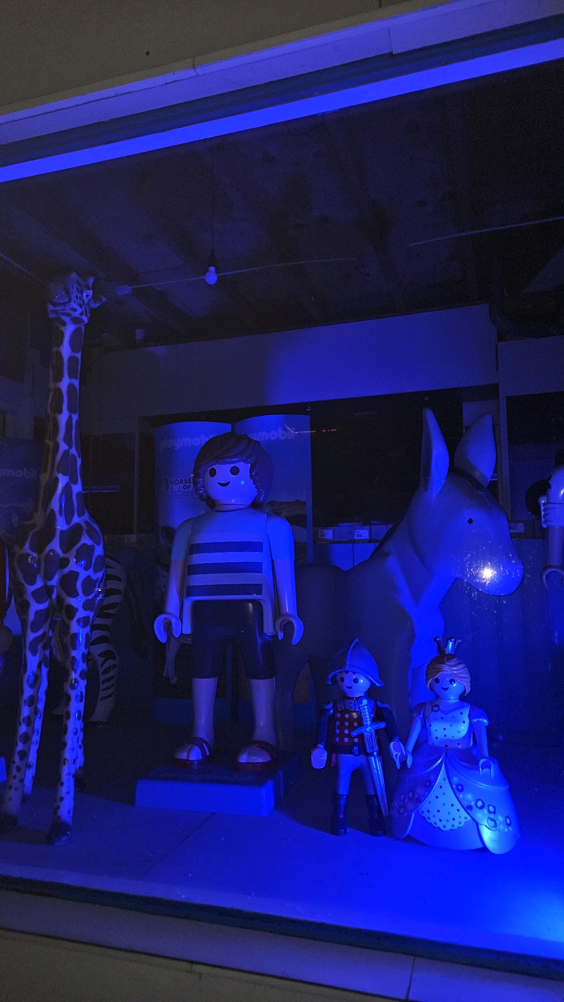 The image shows a display of large Playmobil figures under blue lighting. In the foreground, a giraffe figure stands on the left. To its right is a large figure of a boy with brown hair, wearing a striped shirt and shorts. Behind the boy is a large figure of a horse. In front of the horse, to the right of the boy, are two smaller figures: a knight in armor holding a sword and a princess in a blue dress and crown. The background includes what appears to be advertising banners for "Playmobil" and "Horses of the Sun." The display is set up in what appears to be a commercial or exhibition space, possibly a storefront window. The scene is illuminated with a strong blue light, casting shadows and giving the figures a slightly surreal appearance. The specific location in the city of Breda, Netherlands is not discernible from the image itself.