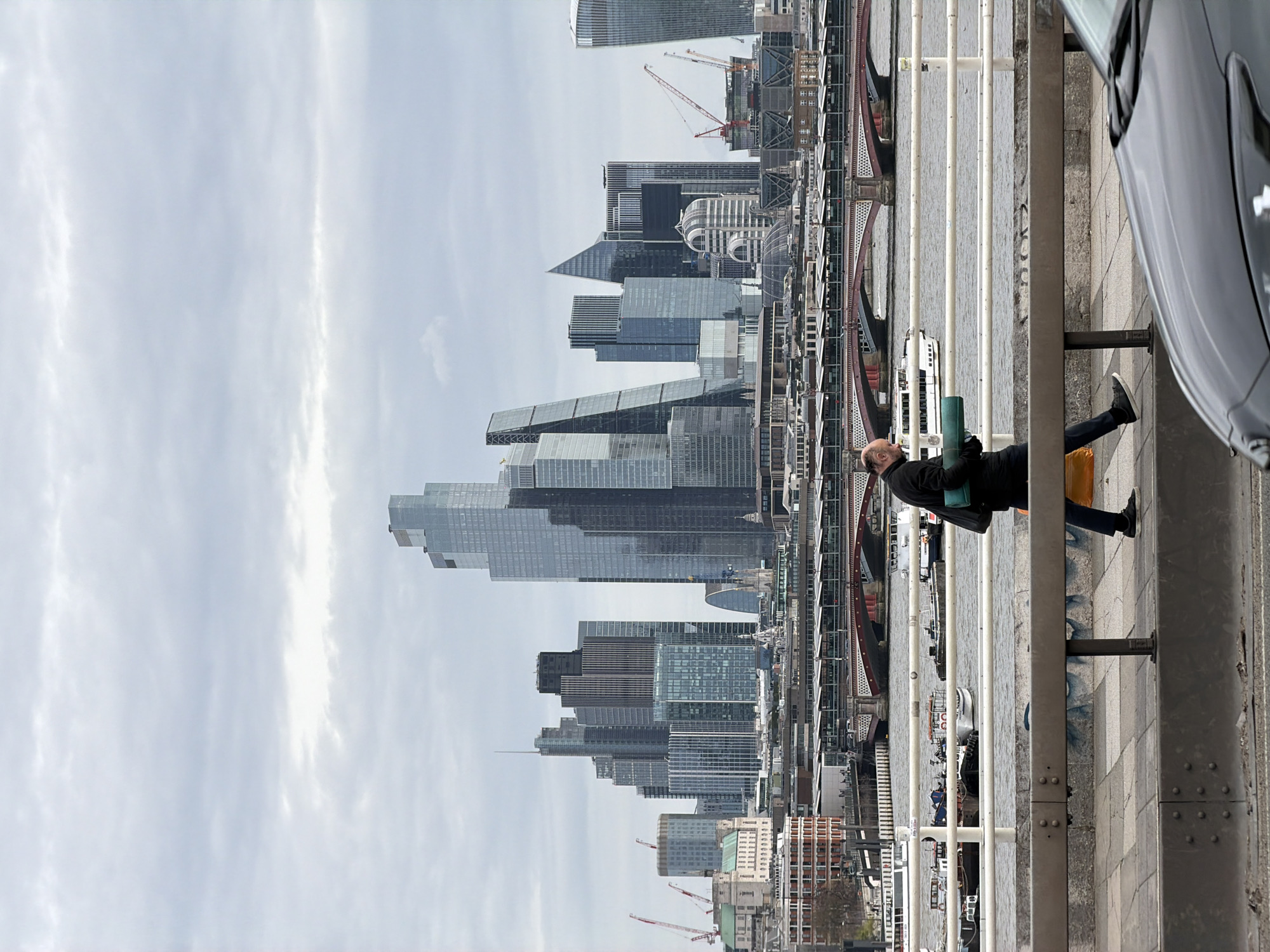 The image is an exterior shot taken during daylight in London, United Kingdom. The vantage point is elevated, possibly from a bridge or high walkway, looking towards the city skyline.
The foreground features a pedestrian area with a metallic railing. A person, possibly male, dressed in dark clothing, leans against the railing. A dark-colored car is partially visible on the far right, suggesting a road is adjacent to the walkway.
The middle ground showcases the River Thames with boats and the distinctive architecture of London. The skyline features modern skyscrapers, including the "Walkie Talkie" building and others under construction (cranes are visible).
The sky is overcast with a diffuse cloud cover, providing soft, ambient light. The scene suggests an urban environment on a cloudy day.