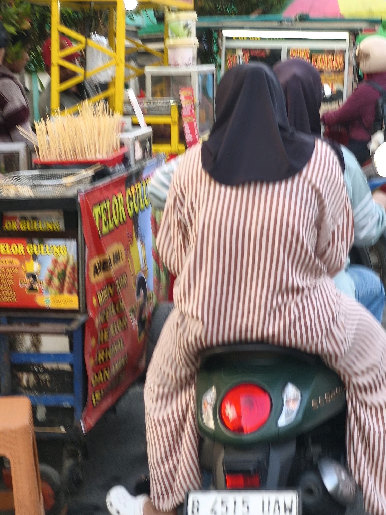 The image depicts a street scene in Satriamekar, Indonesia, likely a market or roadside stall area. The focus is on a scooter with two individuals, viewed from behind. One person is wearing a black hijab and a brown and white striped shirt. The other individual, seated behind, is wearing a light blue shirt. They are both on a green scooter. In the foreground, a red banner with yellow lettering that reads "TELOR GULUNG" (rolled egg) is visible. To the left, a display of food skewers is arranged in a red tray, with additional food advertisements displayed on a blue shelf unit. A person in a maroon shirt and a light-colored helmet is also partially visible on the right side of the frame, suggesting other vendors or customers in the vicinity. The environment appears to be outdoors, with foliage and structures suggestive of a bustling urban or semi-urban area.
