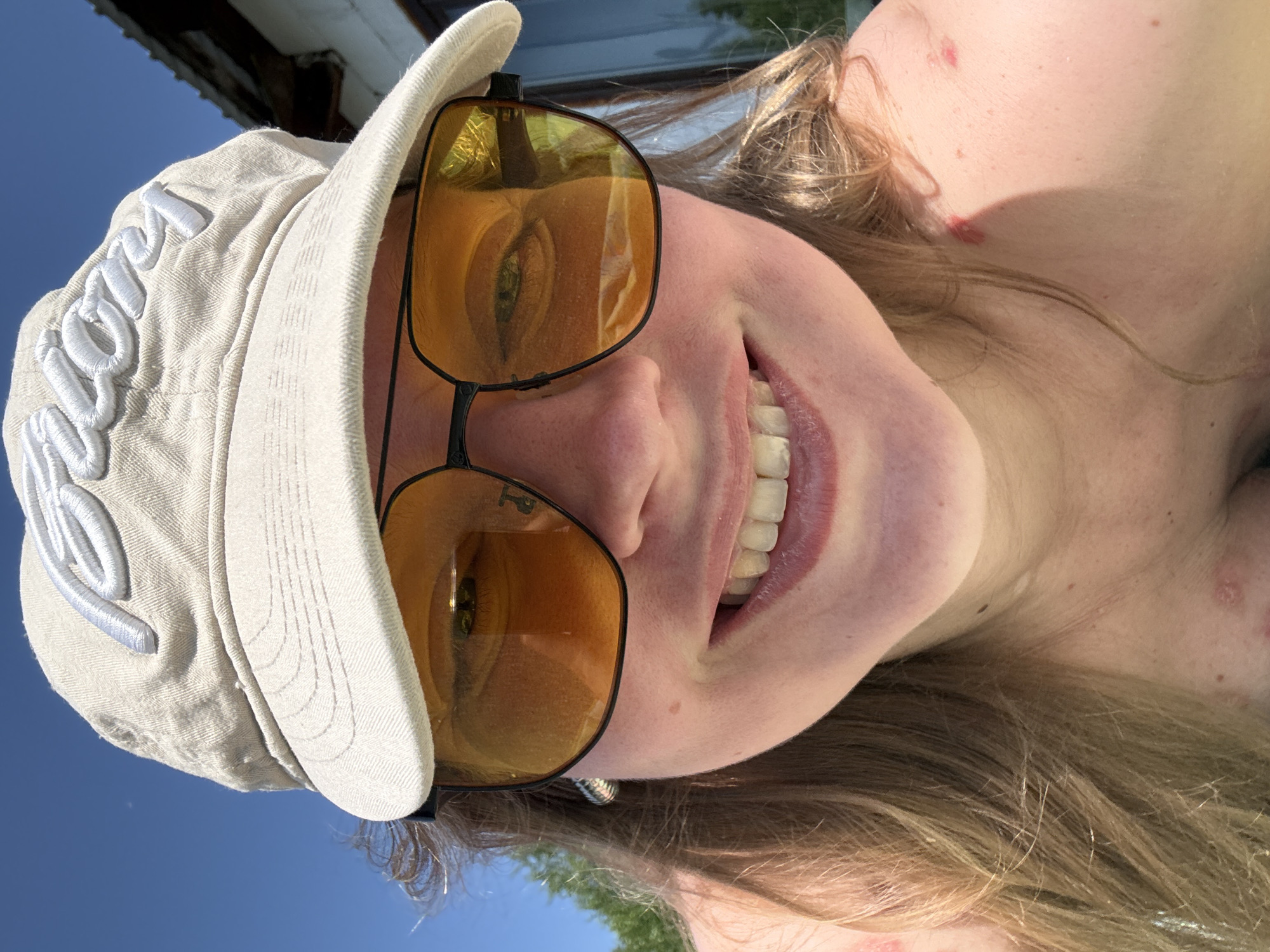 A woman with light skin and blonde hair smiles at the camera, wearing a beige baseball cap with embroidered lettering and yellow-tinted sunglasses. Her teeth are visible in a wide smile. A small portion of a building is visible in the background, along with a patch of blue sky and some green foliage. The location and city are unknown, despite the prompt mentioning Izhevsk, Russia.