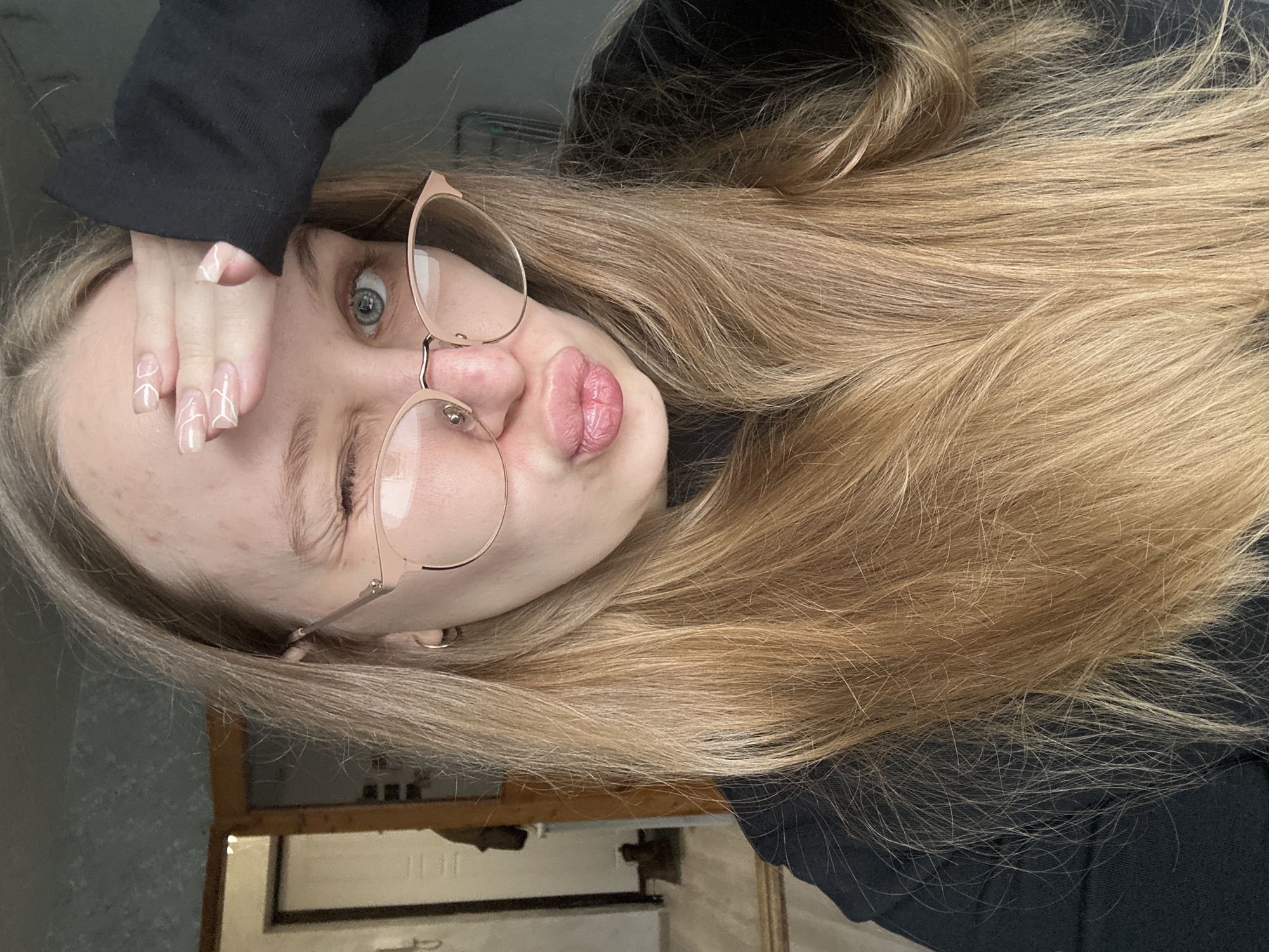 This close-up selfie, rotated 90 degrees counter-clockwise, features a young female with long, light blonde or light brown hair. She is wearing thin, rose gold-framed glasses and a black long-sleeved top. Her expression is playful, characterized by puckered lips often referred to as a "duck face" and a wink with her visible right eye, which is blue or green and looks directly at the camera.
Her left hand is positioned over her forehead, partially obscuring her left eye. Her fingers, which have long, clear or nude-colored nails, are decorated with a distinctive white abstract line pattern. Minor blemishes are visible on her forehead. The background is mostly dark and out of focus, suggesting an indoor setting, possibly a room in Chaikovsky, Russia. The lighting appears natural, consistent with daytime. There is no visible text in the image.