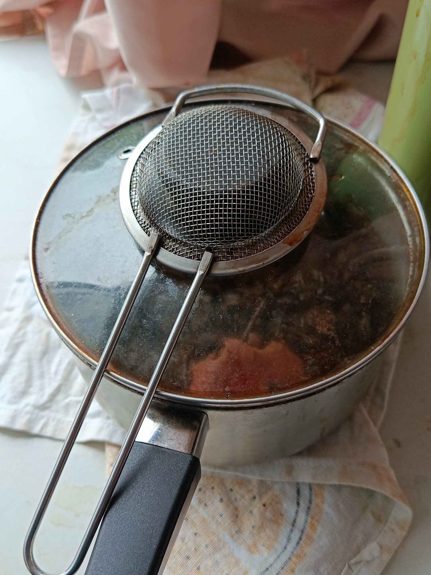 The image shows a close-up, top-down view of a metal pot with a glass lid, a mesh strainer sitting on top of the lid, and a black handle. The pot appears to contain a reddish-brown liquid with some solid pieces. A white cloth with a yellow and blue patterned border is underneath the pot. In the background, out of focus, are pink fabric and a green bottle. There is no visible indication of the city of Halbe, Germany in the image.