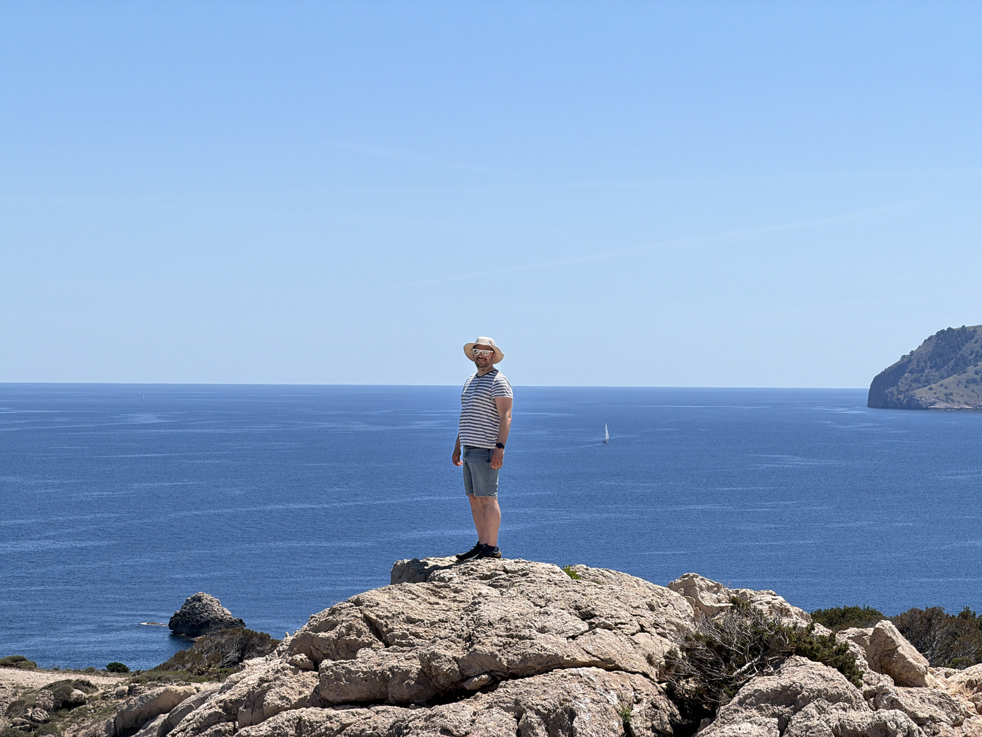 A man stands on a rocky outcrop overlooking the sea in Capdepera, Spain. He is wearing a striped shirt, denim shorts, a hat, and glasses. The sea is blue and there is a small boat in the distance. The sky is clear blue. In the background, there is a land formation. The man is standing still.