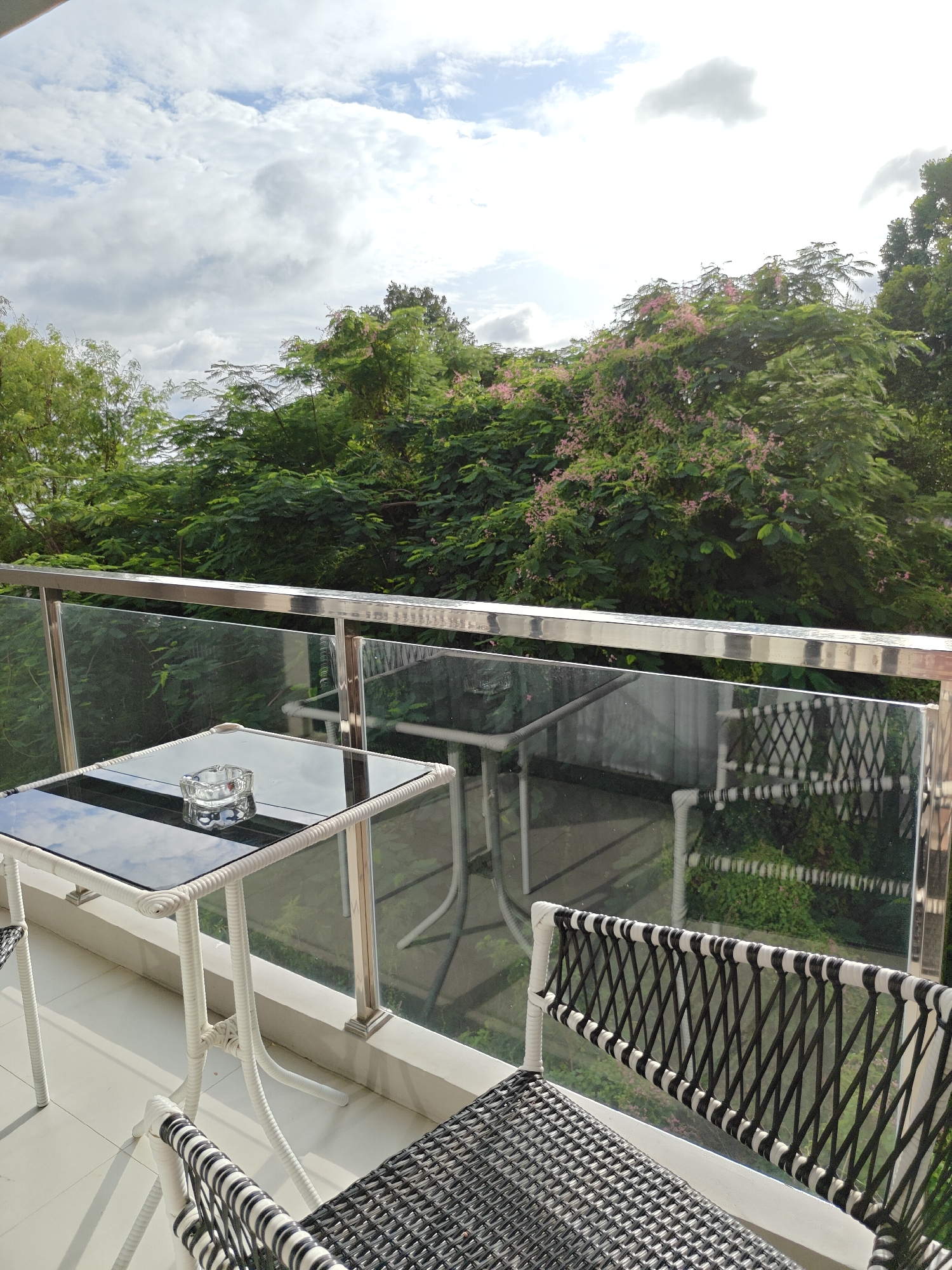A high-angle, medium shot captures a balcony in Pattaya City, Thailand, featuring a small white rattan table with a glass top. An ashtray sits on the table. Two chairs with black and white woven seats and backs are visible, one in the foreground and another partially obscured by the table. A reflective glass railing separates the balcony from an outdoor area with lush green trees, some bearing small pink flowers. The sky above is partly cloudy. The overall impression is that of a serene outdoor seating area.