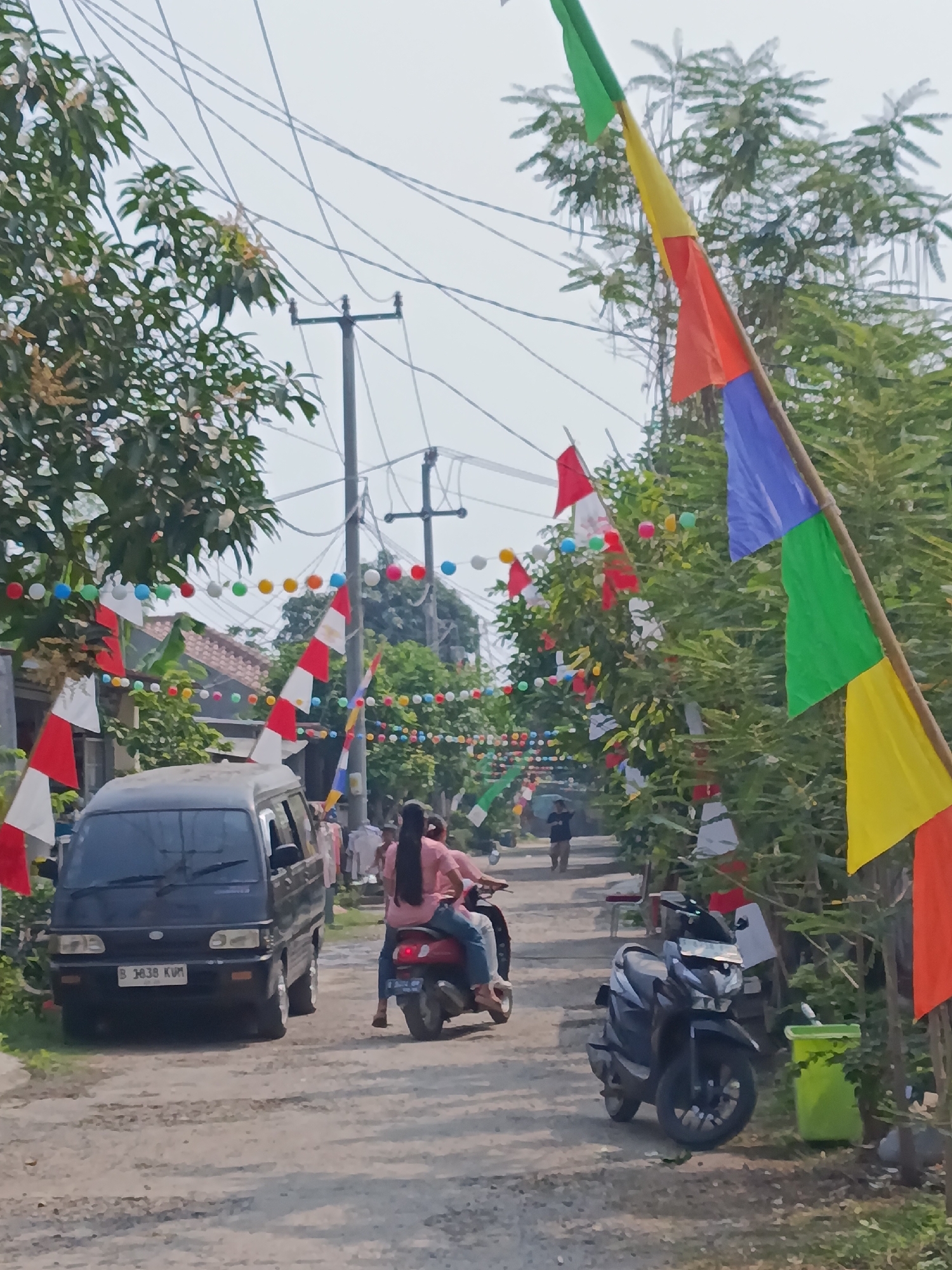The image depicts a street scene in Cariu, Indonesia, decorated with Indonesian flags and colorful pennants strung across the road. On the left side of the unpaved road, a dark-colored van is parked. To the right of the van, a person is riding on the back of a red scooter, with another person in front. Further down the road, a black motorcycle is parked, and a person is walking in the distance. Trees and vegetation line both sides of the street. The sky is overcast, and power lines are visible overhead.