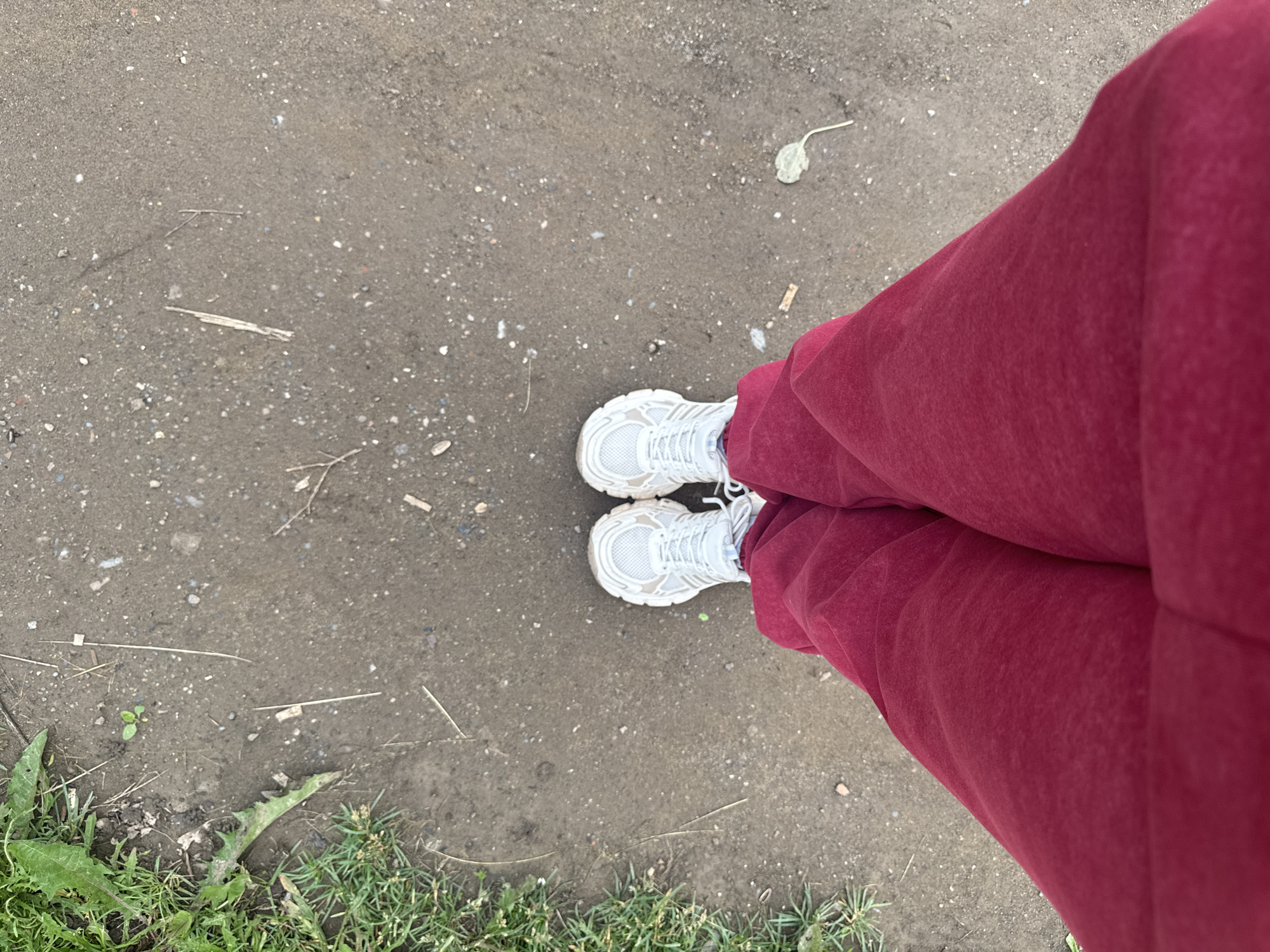 The image shows a pair of feet wearing white sneakers and red sweatpants, standing on a dirt path. To the left of the path is a strip of green grass and some weeds. The path is composed of dirt, small stones, and debris. The image appears to have been taken outdoors in Chastyye, Russia. The perspective is from above, looking down at the person's feet.