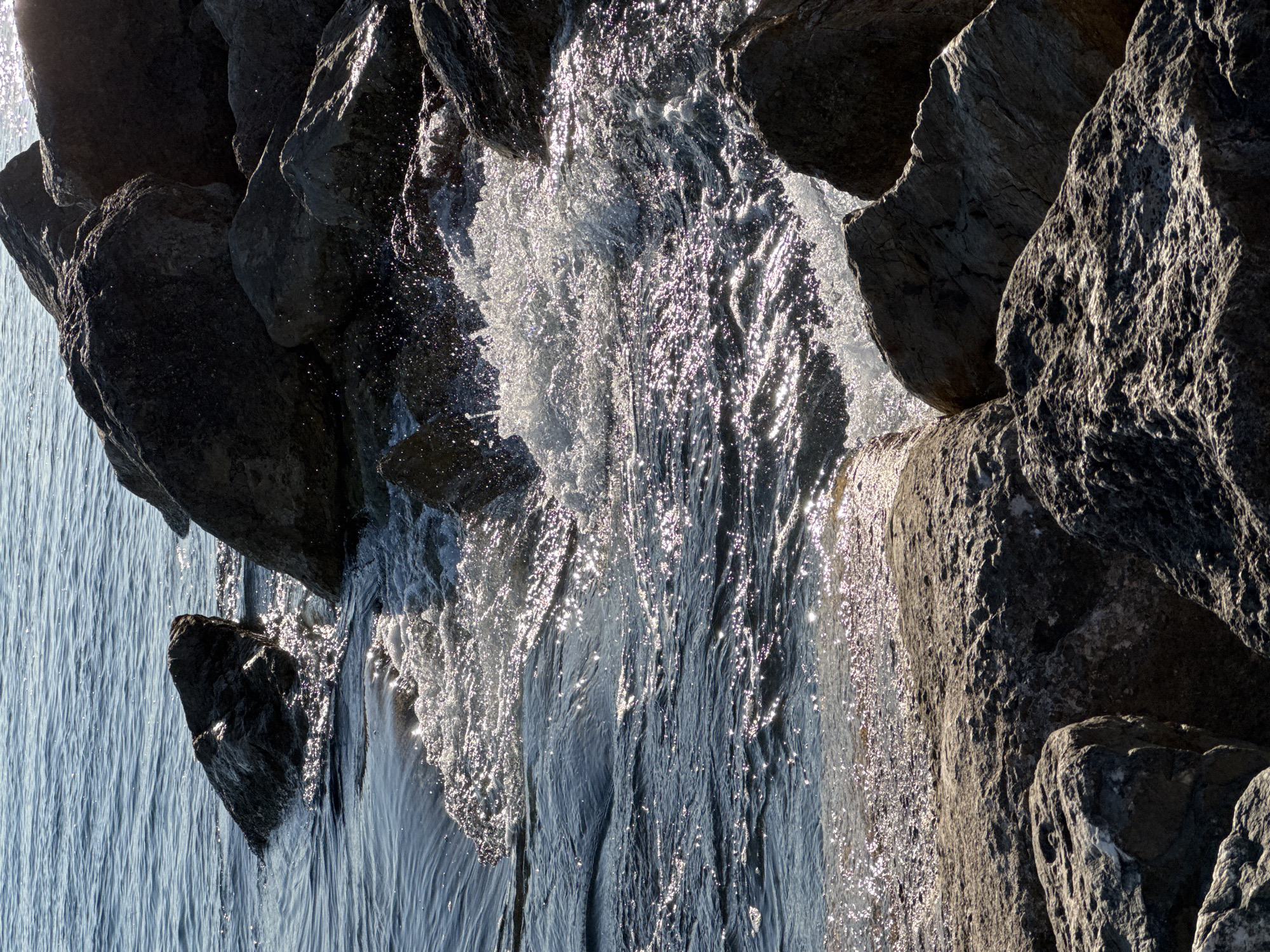 A close-up, sunlit view of waves crashing against large rocks on the coast of Alimos, Greece. The water is a deep blue, sparkling with highlights from the sun. White foam and spray erupt where the waves meet the rocks. The rocks themselves are varied in size and texture, with some appearing smooth and others rough and weathered. The scene captures the dynamic interaction between the sea and the rocky shoreline.