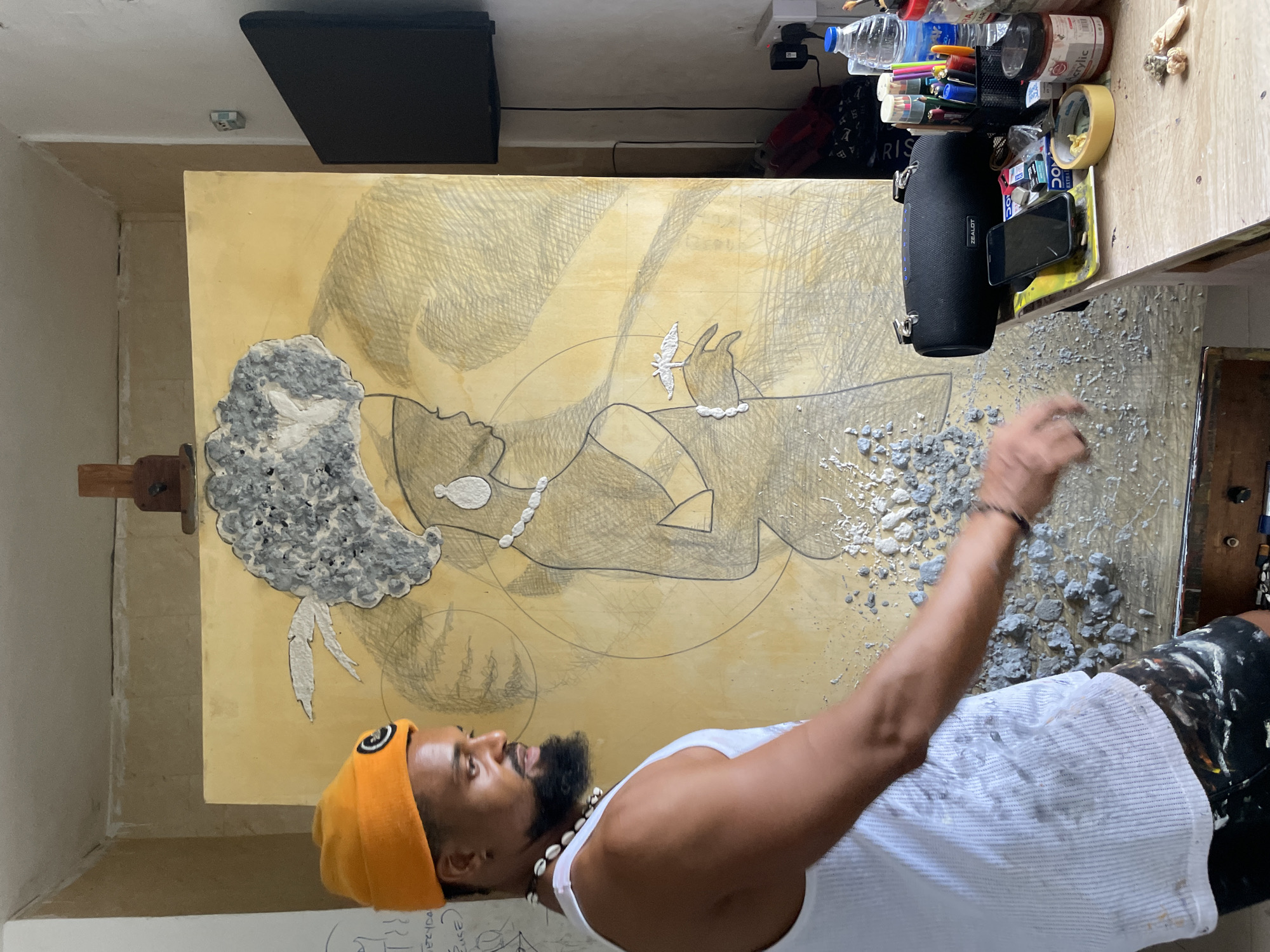 A medium-angle shot depicts a man working on a piece of art within an indoor setting. The man, wearing a white tank top, an orange beanie, and a necklace, stands to the right of the artwork, which is an illustrative piece on a tan-colored canvas mounted on an easel. The illustration includes a side profile of a person and decorative abstract patterns. The man's right arm is extended towards the canvas, suggesting he is in the process of creating or adjusting the artwork.
The surrounding environment appears to be an artist's workspace, as indicated by the various art supplies on a table to the upper right. The table holds water bottles, pens, paint containers, masking tape, and a black speaker. A black rectangle hangs on the wall above the artwork, possibly a monitor.
The location is Ado, Nigeria, implied through external context or metadata not directly evident from the image.