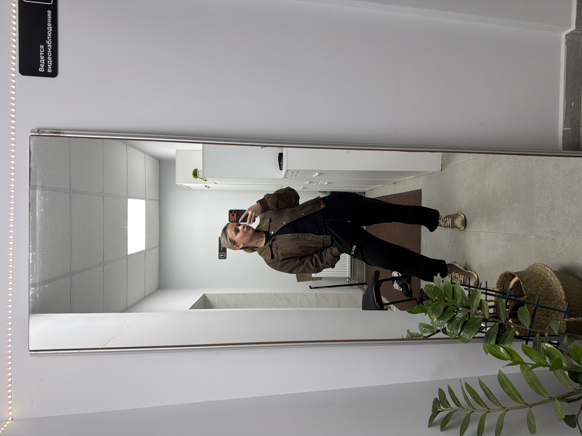 A young woman is taking a selfie in a mirror in a room. She is wearing a brown jacket, a black top, black pants, and tan sneakers. The room appears to be a restroom with white tile walls and a tiled floor. The reflection reveals lockers and a bright overhead light. A plant sits in front of the mirror. A sign in Russian is affixed near the top of the mirror. The location is Sarapul, Russia.