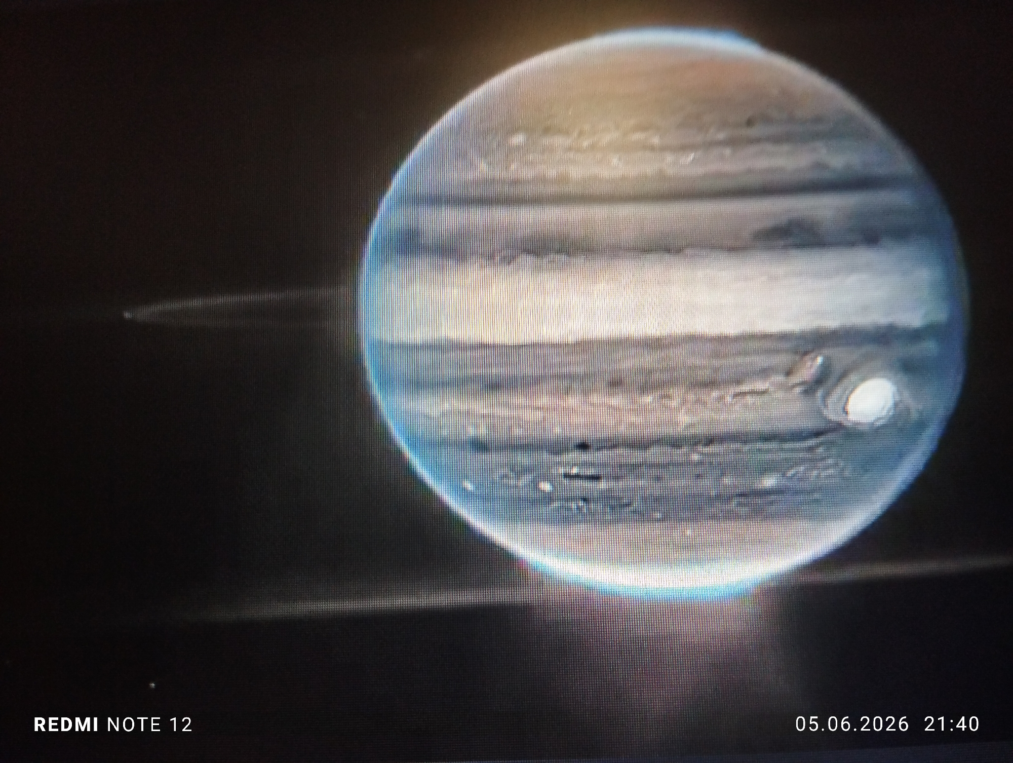 The image displays a photograph of a digital screen showing a detailed astronomical image of the planet Jupiter. Jupiter is rendered with its characteristic horizontal cloud bands in shades of blue, white, brown, and orange, and a prominent bright white, oval-shaped atmospheric feature is visible on its lower right quadrant. Faint, ethereal arc-like structures are discernible to the left of the planetary disk. The entire image exhibits a visible vertical pixel grid pattern, confirming it is a photograph taken of a digital display. In the bottom left corner, the text "REDMI NOTE 12" indicates the make and model of the device used to capture the photograph of the screen. In the bottom right corner, a timestamp "05.06.2026 21:40" specifies the date and time when the photograph of the screen was taken. There are no people present. The environment is dark, typical of a screen displaying an astronomical subject. No visual cues within the image provide any information regarding the location in the city of Burgas, Bulgaria.