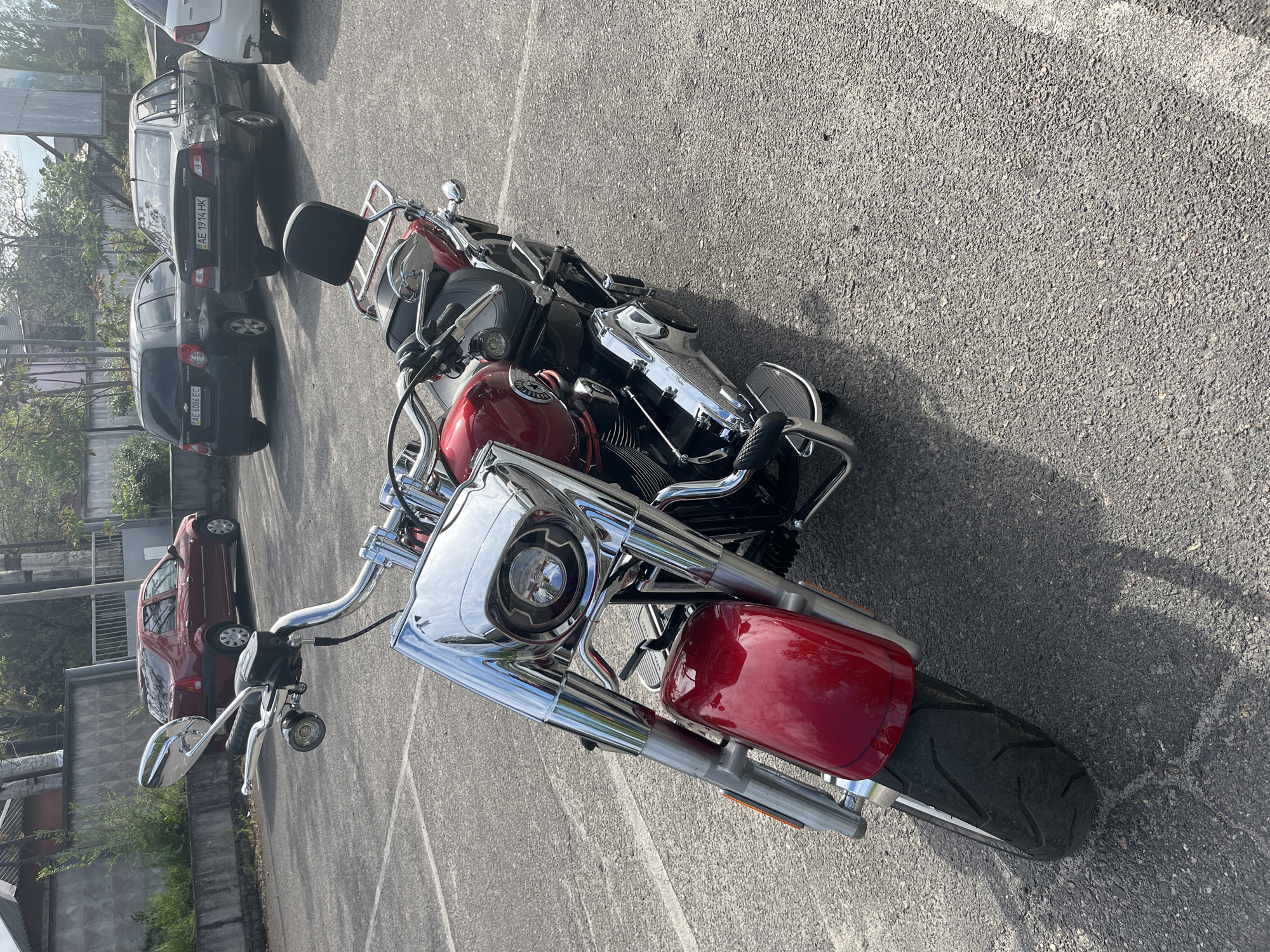 The image shows a red and chrome motorcycle parked on an asphalt surface. In the background, several cars are parked, including a white car, a dark gray car, and a red car. The license plates of the gray car read "AE 1914 HK" and "AE 6866 EC". The photo appears to have been taken outdoors in a parking area. The location is Kryvyi Rih, Ukraine.