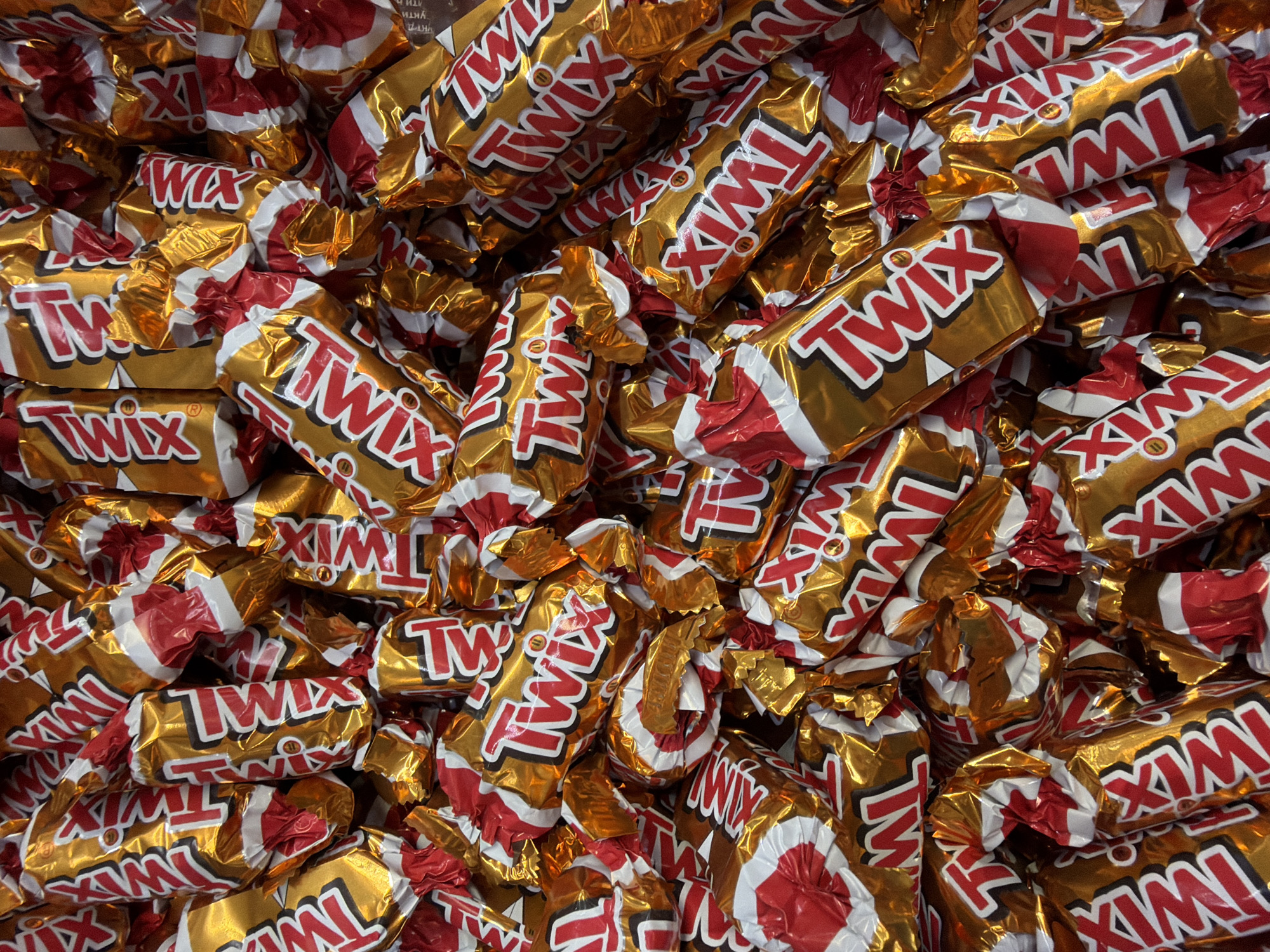 This is a close-up, overhead shot of a large pile of individually wrapped Twix candy bars. The wrappers are shiny gold with red and white lettering that says "Twix". The candy bars are oriented in various directions, creating a dense, overlapping mass. The lighting is bright and even, highlighting the reflective surfaces of the wrappers. There are no people visible in the image. The context of Sokilnyky, Ukraine is not apparent from the visual information provided.