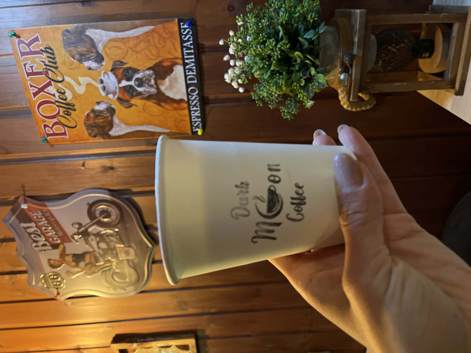 A hand holds a white coffee cup with "Dark Moon Coffee" printed on it in front of a wooden wall. In the background, a "Boxer Coffee Club" sign, a small potted plant, and a "Best Garage" motorcycle sign are visible. There's no indication of a location in Petropavlovsk-Kamchatsky.