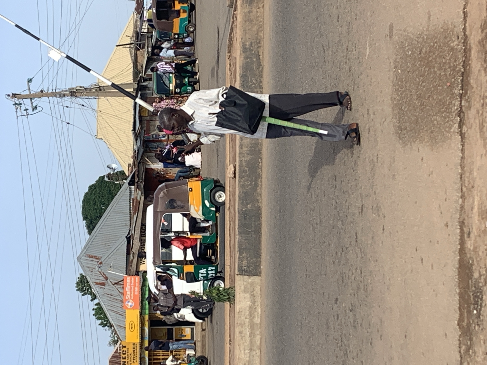 The image depicts a street scene in Gura Top, Nigeria, with a man standing on a paved road, holding a black bag and a green and white striped object that appears to be a walking stick or cane. He is wearing a striped long-sleeved shirt and dark trousers, with sandals on his feet. The man is looking to his left. In the background, there are several "tuk-tuks" (auto-rickshaws), one of which is white and has a number plate that reads "PTA 317". Other vehicles, possibly also tuk-tuks, are visible, including a green and yellow one. Buildings line the street, with corrugated metal roofs. A power pole with numerous wires stands prominently on the right side of the image, supporting a black and white striped barrier that extends diagonally upwards. Some people are visible near the buildings and in the vehicles, suggesting a bustling urban environment. The sky is clear and bright, indicating daylight. The foreground shows the asphalt road with a concrete curb.