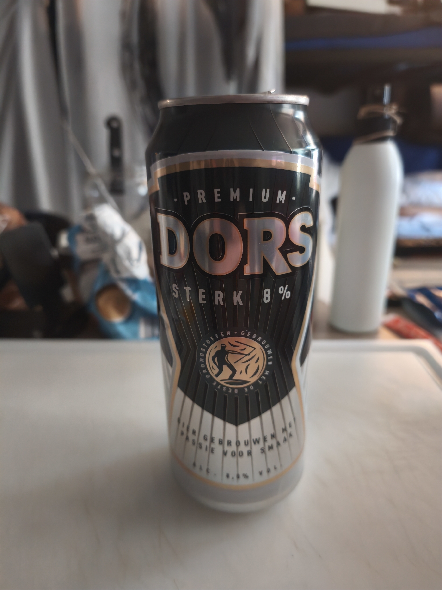 A close-up, slightly angled, medium shot shows a dark metal beer can with a metallic gold and silver label. The can is positioned vertically and is the primary subject. The label displays the brand name "DORS" in large, bold, metallic gold letters. Above "DORS", "PREMIUM" is written in smaller, metallic gold letters. Below "DORS", "STERK 8%" is also visible in metallic gold. A circular emblem with a silhouetted figure is present on the lower half of the label. Text on the can is predominantly in Dutch, including "BROUWEN MET PASSIE VOOR SMAAK" and "ALC. 8.0% VOL". The background is blurred, showing a light-colored surface that appears to be a table or countertop in the foreground, with a white bottle and other indistinct objects in the midground and background. The overall lighting is dim and appears to be indoors. No specific identifiable landmarks or features within the city of Assen, Netherlands, are visible in this image.