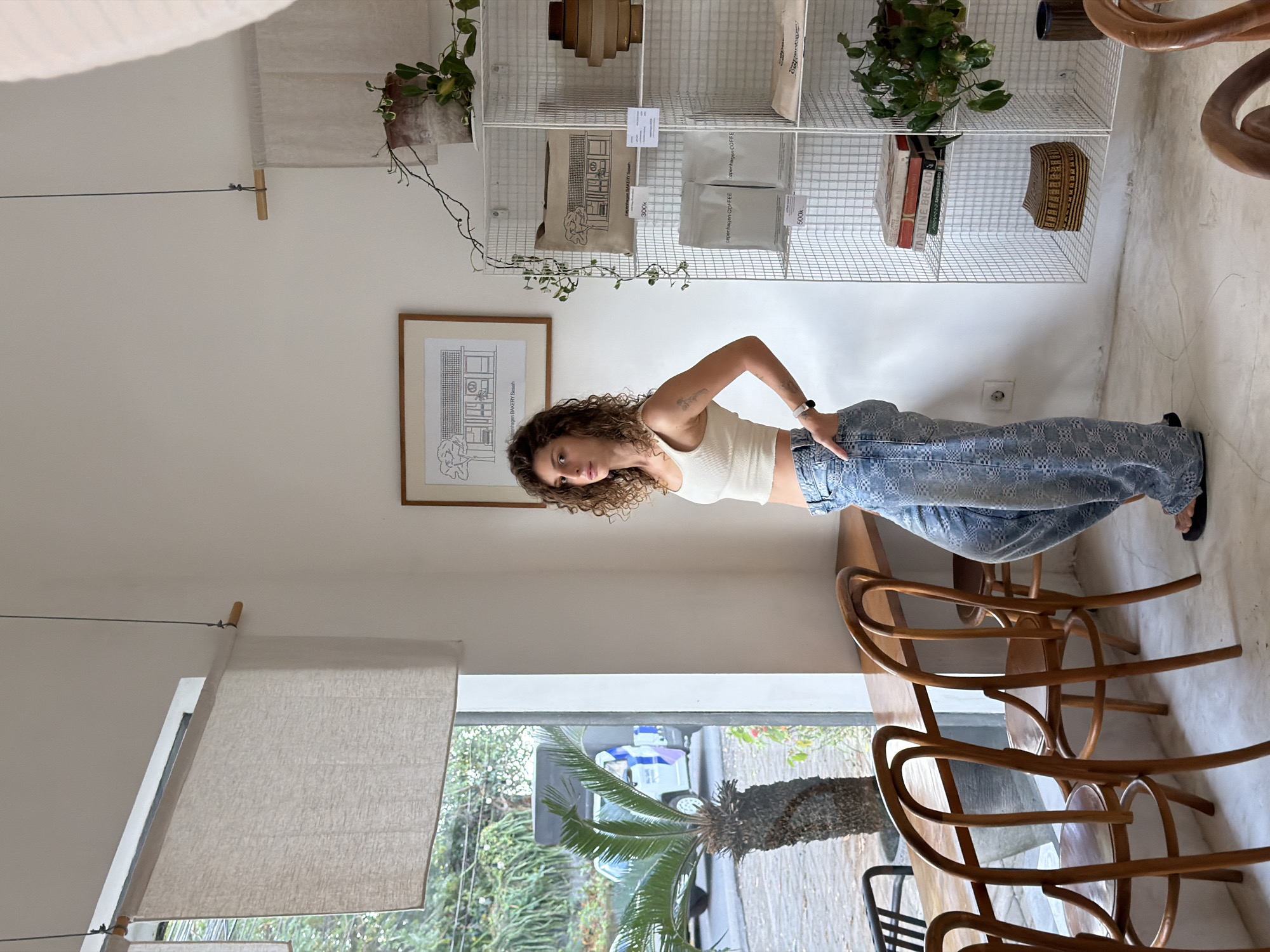 A woman with curly brown hair stands in a cafe in Mengwi, Indonesia, leaning against a wooden table. She is wearing a white crop top and wide-leg patterned jeans. The cafe has large windows overlooking a street with palm trees and a white truck. There are wooden chairs around the table and a white wire shelving unit filled with plants, books, and packaged goods. The walls are white, and there is a framed drawing of a building on the wall.