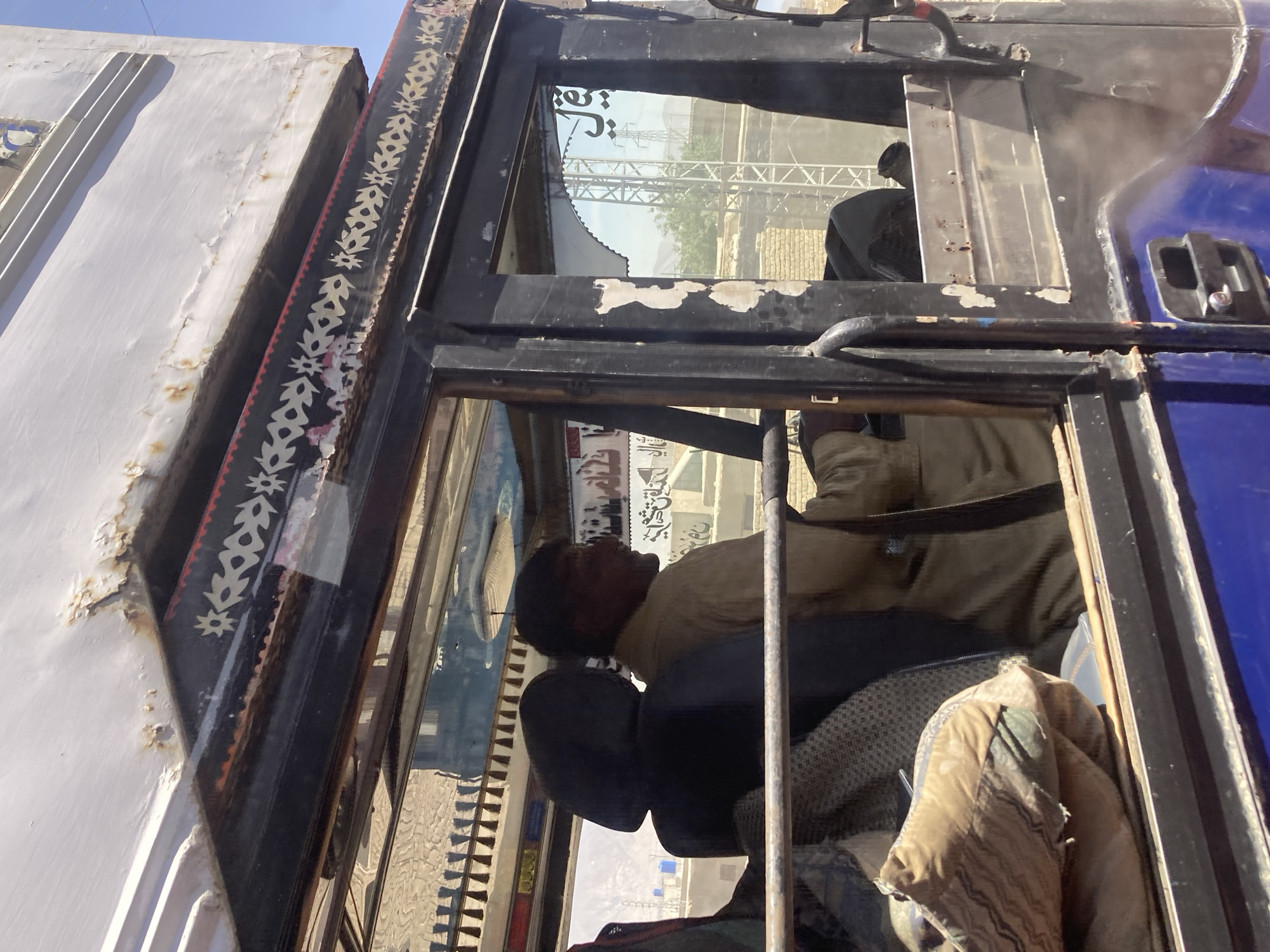 This image offers a close-up, angled view into the cabin of a large vehicle, likely a truck or bus, adorned with distinctive "truck art" styling. A man with short, dark hair is visible inside, resting with his head tilted back, appearing to be asleep or taking a break. He is wearing a light brown or tan-colored shirt.
The vehicle's exterior features a dark-colored frame, intricately decorated with white floral and star-like patterns on a black background, highlighted with red accents. Adjacent to this highly decorated area, a part of the vehicle's body is dark blue, while a separate, taller structure to the left appears white with visible peeling paint and rust.
Through the cabin windows, an outdoor environment under a clear blue sky is visible. The background shows distant arid hills, industrial or building structures, and sparse vegetation, consistent with a dry climate. Inside the cabin, a dark headrest, a patterned seat cover, and a crumpled light brown or greenish fabric item are discernible.
Notable details include stylized text in what appears to be Arabic or Persian script, forming part of the window decorations and visible as faint reflections or embellishments within the cabin. The scene is set during bright daylight hours, indicated by the strong sunlight and clear sky. The decorative elements on the vehicle are characteristic of regional truck art, suggesting the location context of Quetta City Tehsil, Pakistan.
