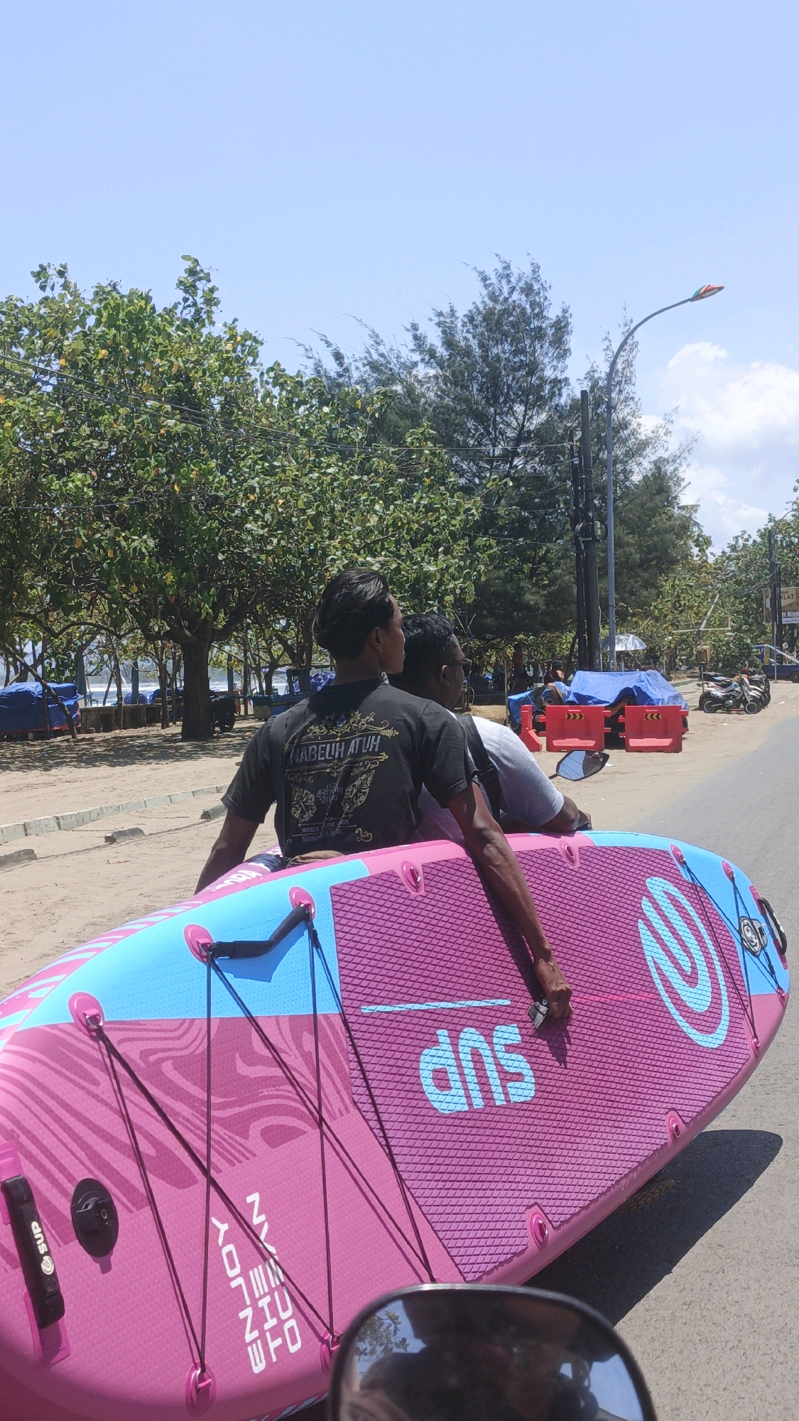 Two men are riding on a motorbike on a street in Desa Pagandaran, Indonesia, carrying a large, pink and blue stand-up paddleboard. The man in front is wearing a black t-shirt with text on the back. The paddleboard is strapped to the back of the motorbike and extends in front of the riders. The street is lined with trees and appears to be near a beach, with beach umbrellas and tents visible in the background. The sky is clear and blue, indicating a sunny day. The overall scene suggests a recreational outing or transport of sports equipment.