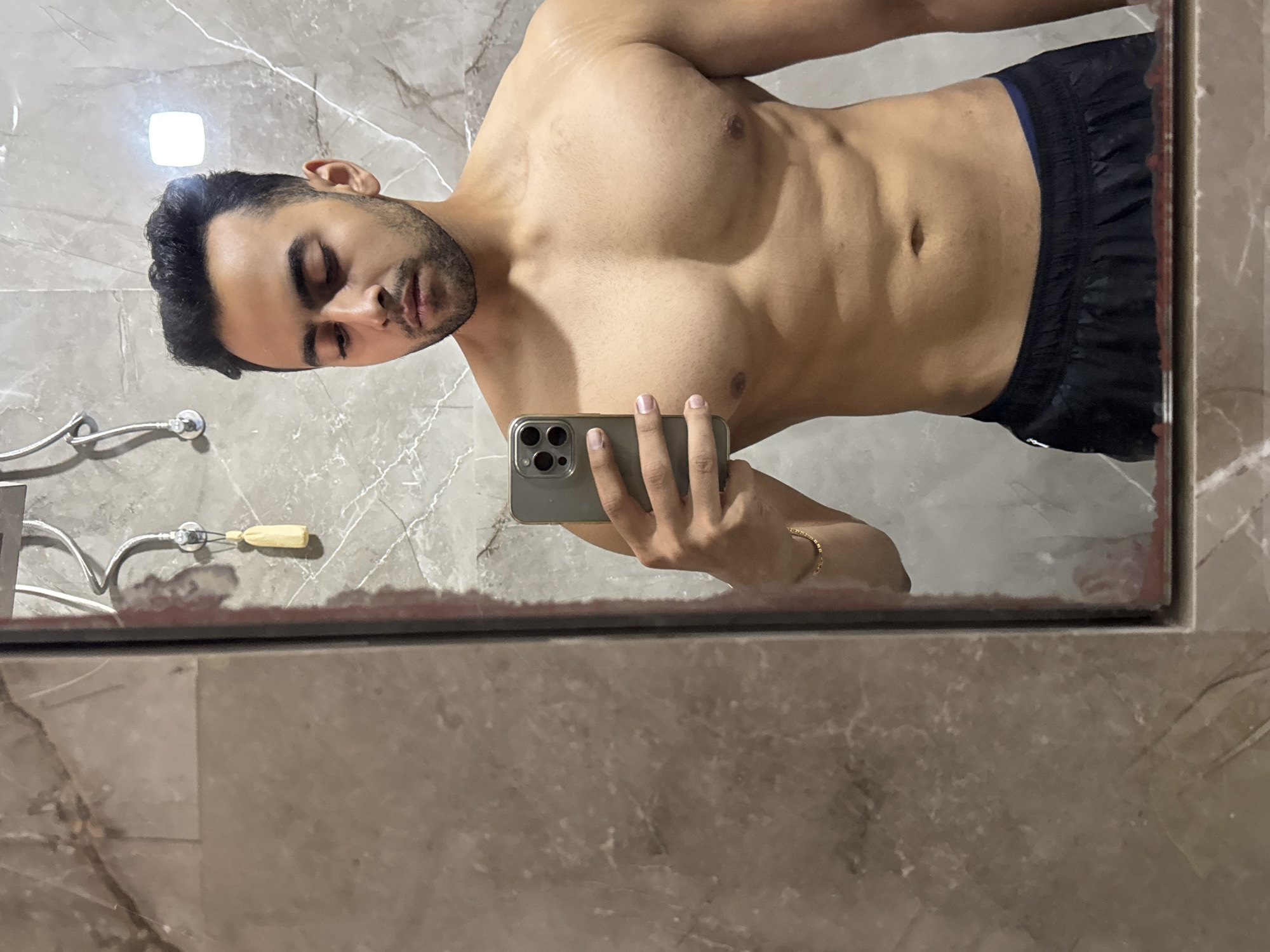 A muscular man is taking a selfie in a bathroom mirror. He is shirtless and wearing dark shorts with a blue waistband. He is holding a gray phone in his right hand. The bathroom has gray marble walls. There is a shower fixture with a tan handle in the upper left of the image. The location is unknown and cannot be determined from the image. The city of Kolkata, India, is not discernible from the image.
