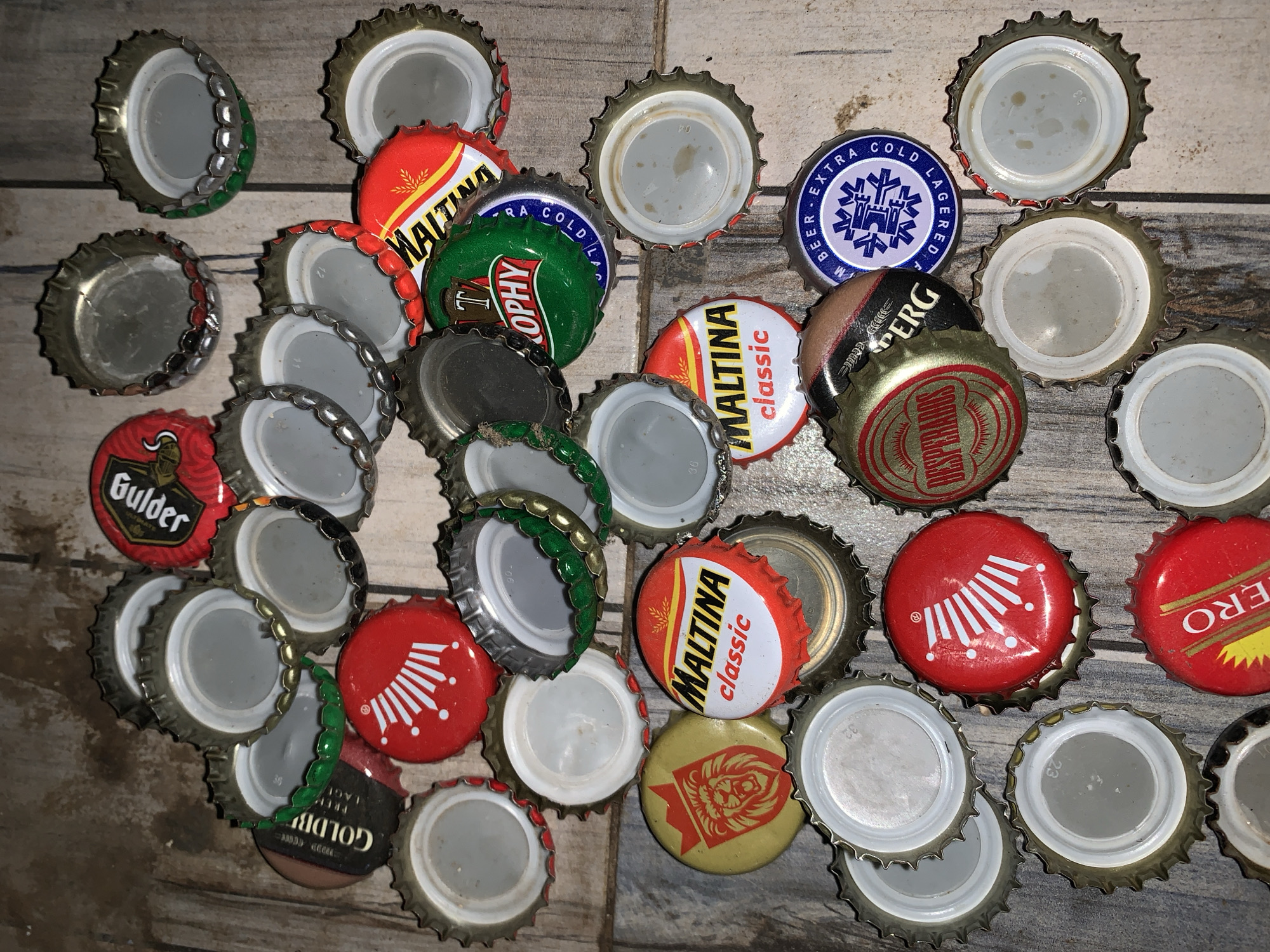 The image shows a collection of approximately 30 bottle caps scattered on a surface resembling a tiled floor with a light-colored, possibly wooden, pattern. The bottle caps vary in color and design, featuring brands like "Gulder", "Maltina Classic", "Trophy", "Desperados", "Goldberg Lager", "Extra Cold Lager", and "ZERO". The caps are arranged randomly, overlapping in places, and some are turned inside up, revealing the plain metal interior.
There is no person in this image.
There is no action happening.
The environment looks like a table.
The image does not provide any information about New Stadium or Nigeria.