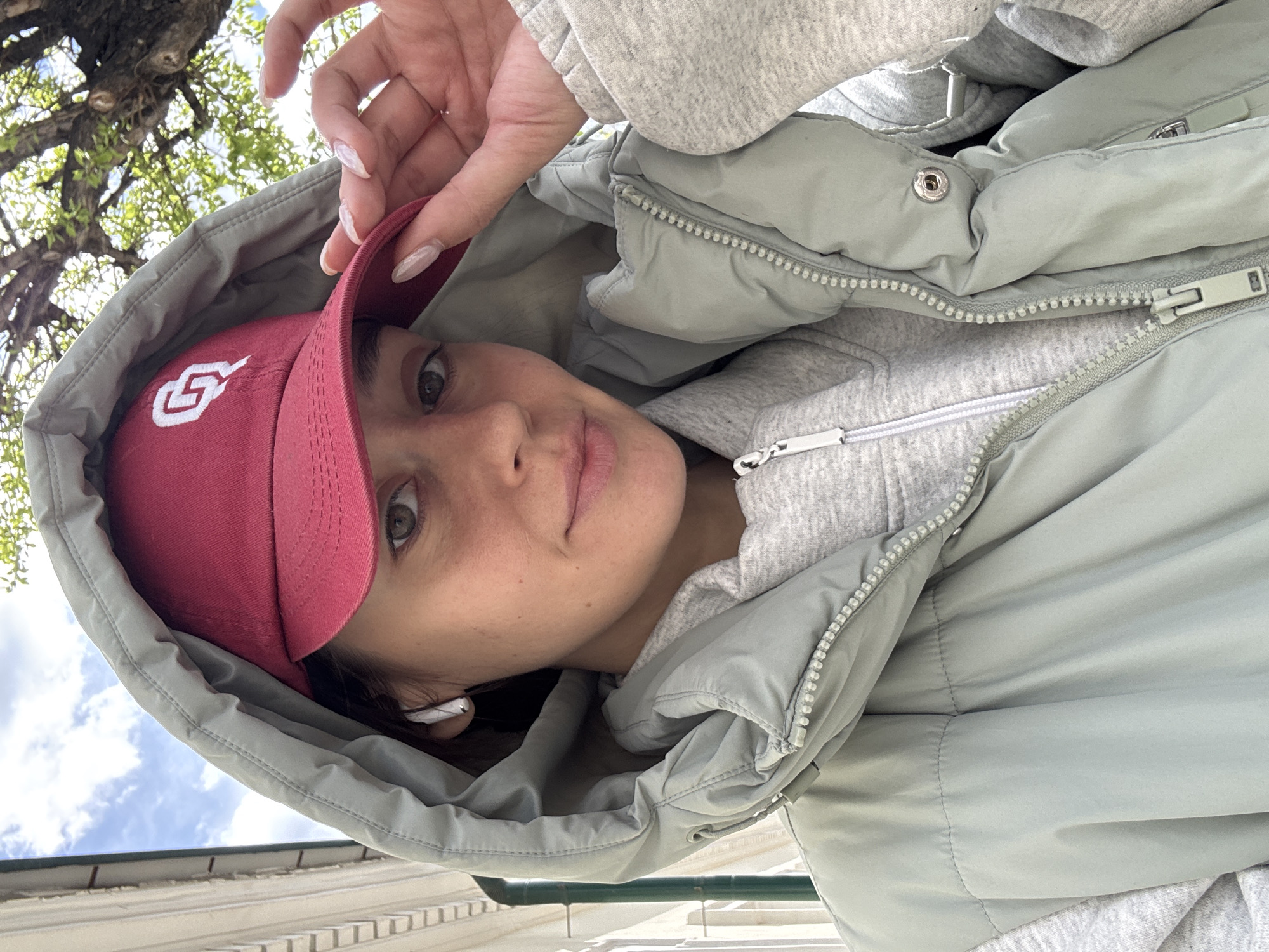 A woman is taking a selfie outdoors. She's wearing a red baseball cap with a white logo, a gray hooded sweatshirt, and a light green puffy jacket with the hood up. Her hand is touching the bill of the cap. A white earbud is visible in her ear. In the background, there are trees and a blue sky with clouds. The exact location in Krasnoyarsk, Russia, cannot be determined from the image.