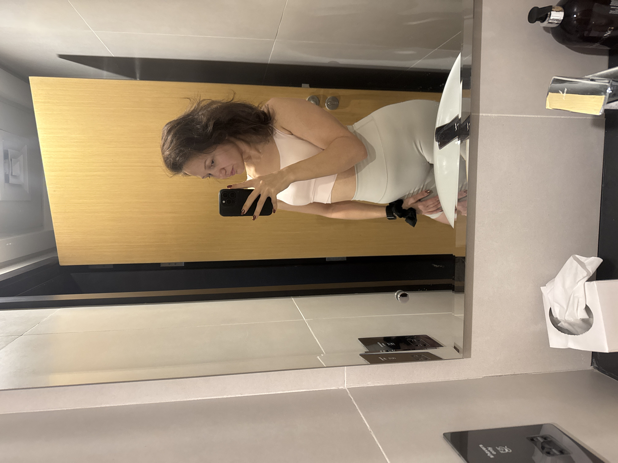 A woman is standing in a bathroom, taking a photo of herself in the mirror with her phone. She is wearing a light pink sports bra and light-colored leggings. The bathroom has a wooden door, light gray tiled walls, and a countertop with a sink and toiletries. The location is Dubai, United Arab Emirates.