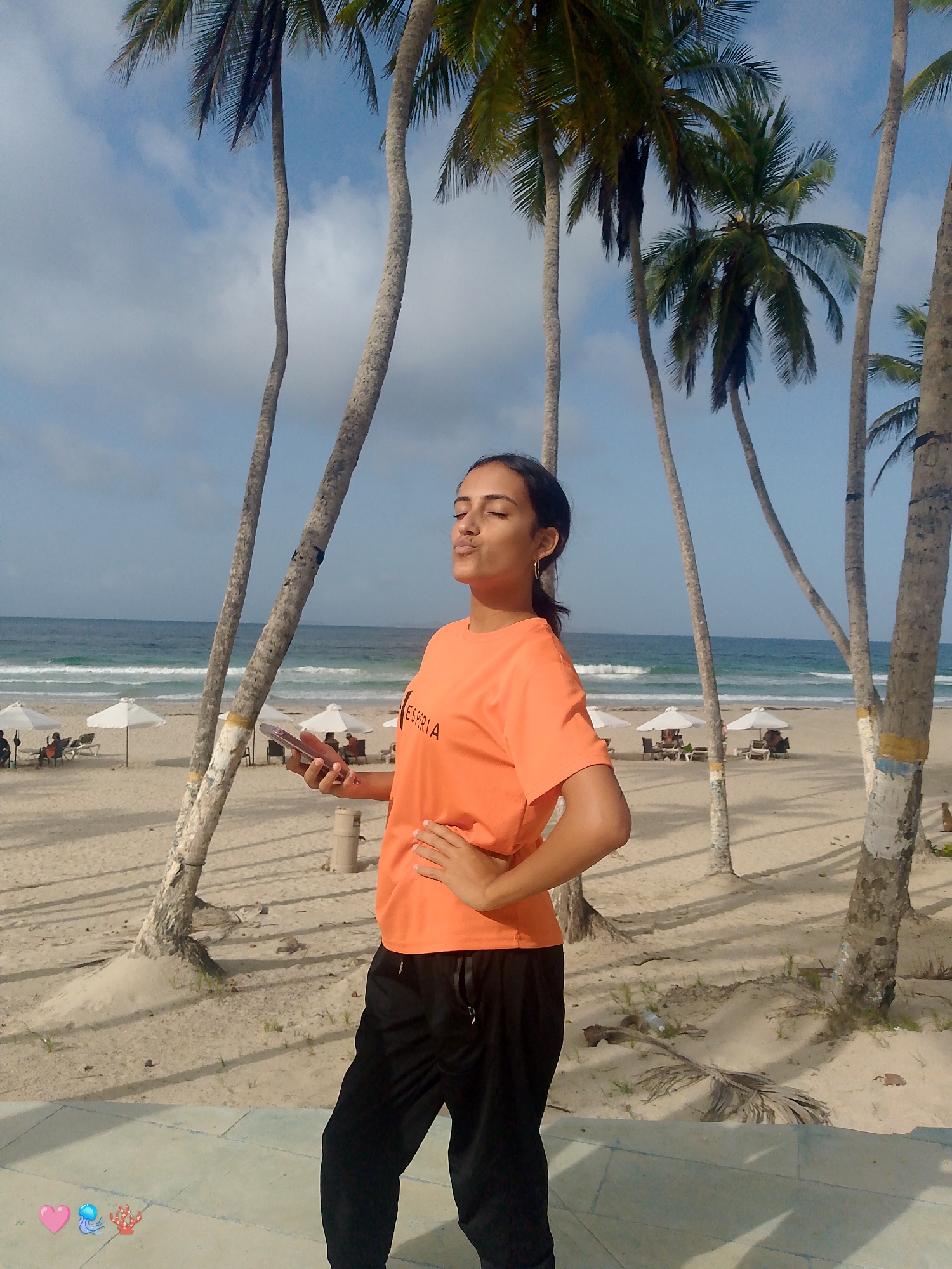 The image shows a young woman standing on a paved area at Playa El Agua, Venezuela, with the beach and ocean in the background. She is wearing an orange t-shirt with the word "ESPRIA" on it and black athletic pants. Her eyes are closed and her lips are pursed as if blowing a kiss, and her right hand is on her hip. She is holding a pink mobile phone in her left hand. Several palm trees are visible around her, and the beach is dotted with white umbrellas and lounge chairs. The ocean is calm with gentle waves. The sky is partly cloudy.