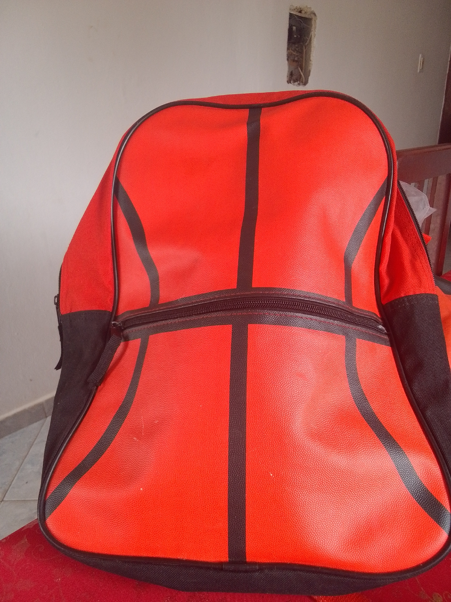 The image shows a red backpack designed to resemble a basketball. The backpack has black lines mimicking the seams of a basketball. It has a zippered opening across the front. The backpack is placed on a red surface, possibly a chair or a table, and is set against a plain, light-colored wall. The setting appears to be indoors, with some indistinct background elements that suggest a utilitarian or storage area. There is no visible text in the image. The lighting is even, suggesting it is daytime but not direct sunlight. The overall impression is of a sports-themed accessory in a domestic or storage environment.