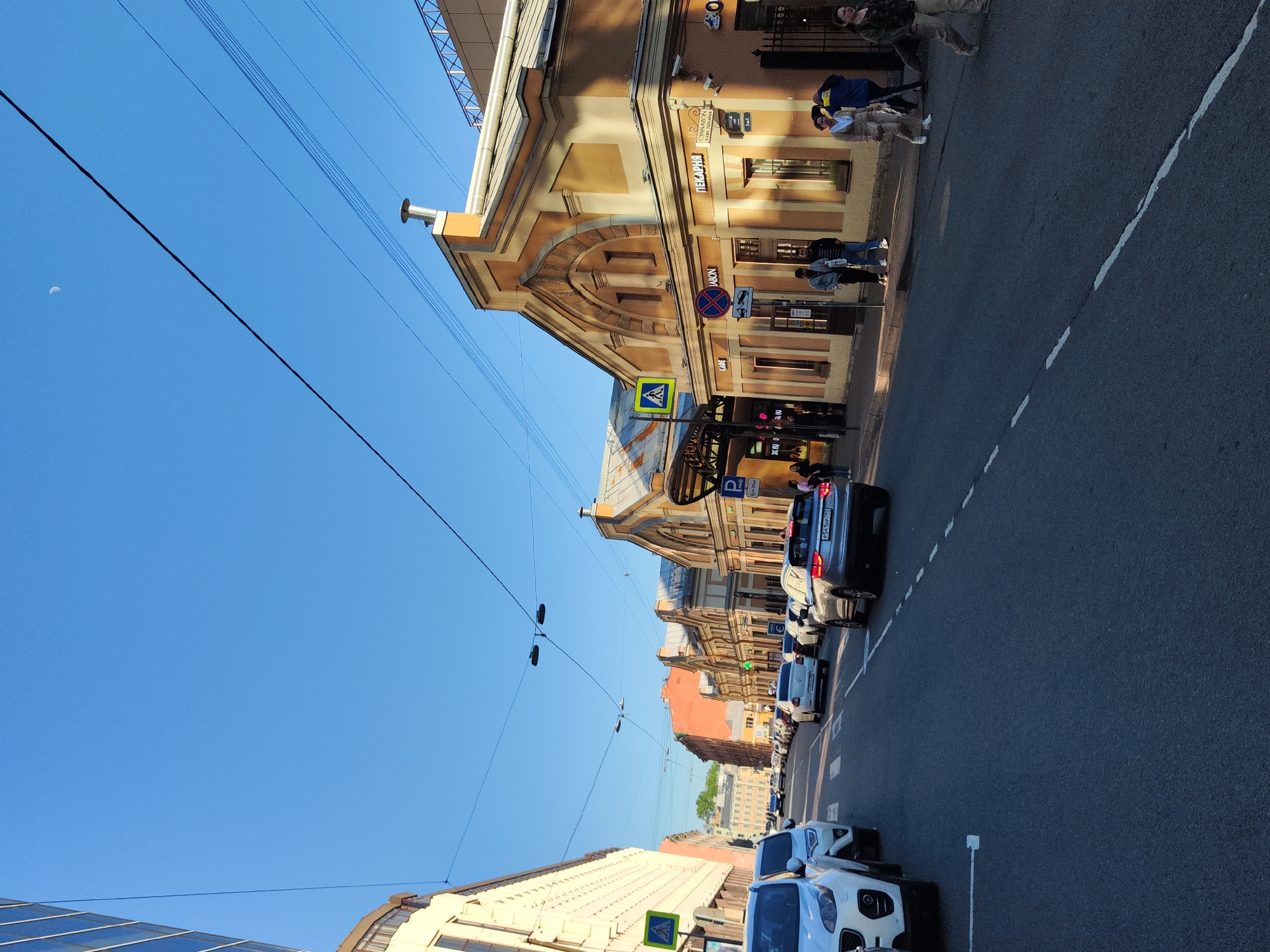 The image shows a street scene in Saint Petersburg, Russia, with various buildings and a clear blue sky. There are several cars parked along the right side of the street, and people are walking on the sidewalk next to the buildings. Utility lines are visible above the street, extending from the left side of the frame towards the right. The buildings have architectural details and signage in Russian. Street signs with crosswalk markings are also visible.