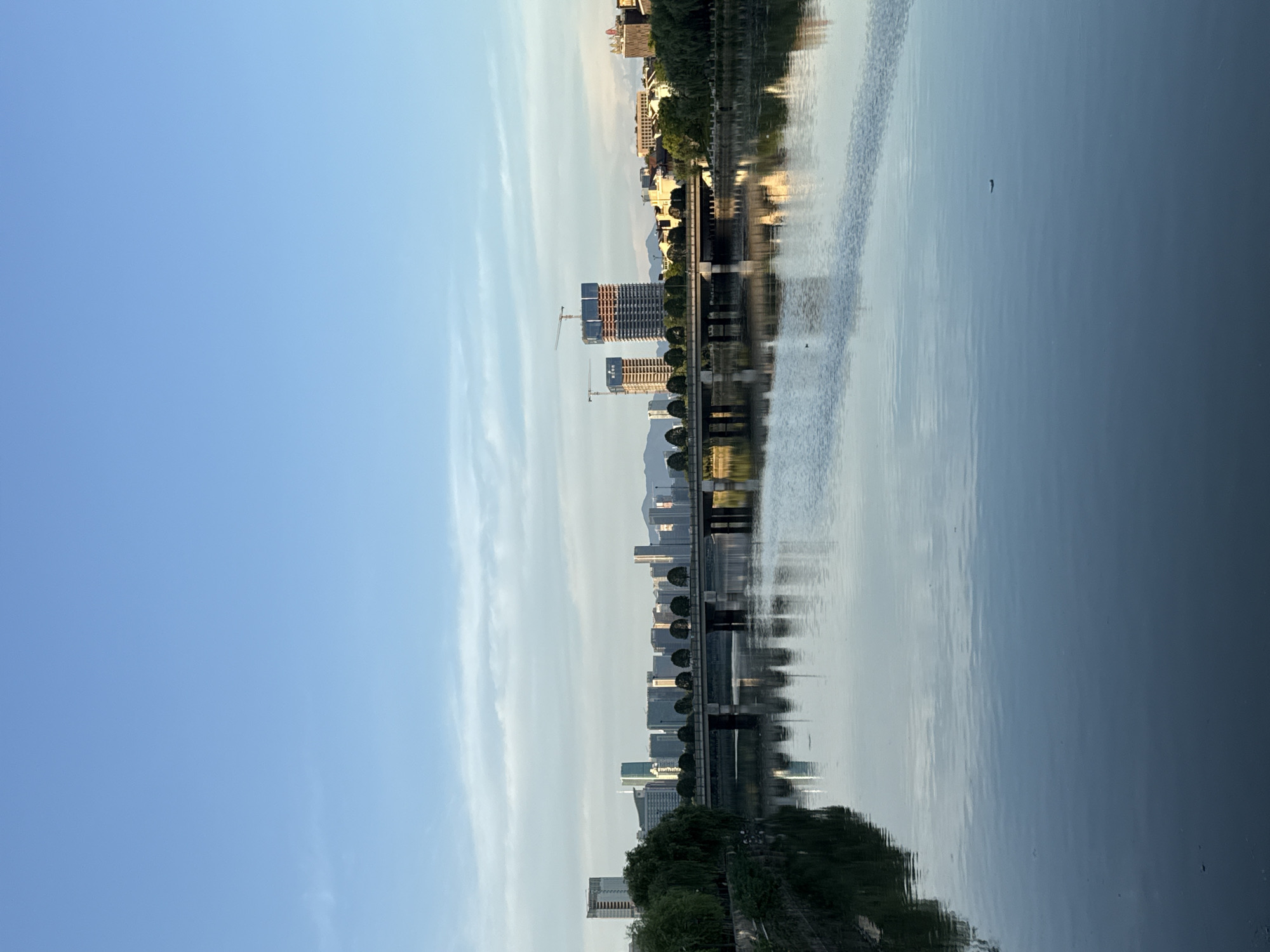 The image shows a wide, calm river in the city of Yiwu, China, reflecting the clear blue sky and a city skyline. A bridge spans the river, with buildings and construction sites visible on the opposite bank. Lush green trees line the riverbanks. The scene appears serene, likely captured during the day.