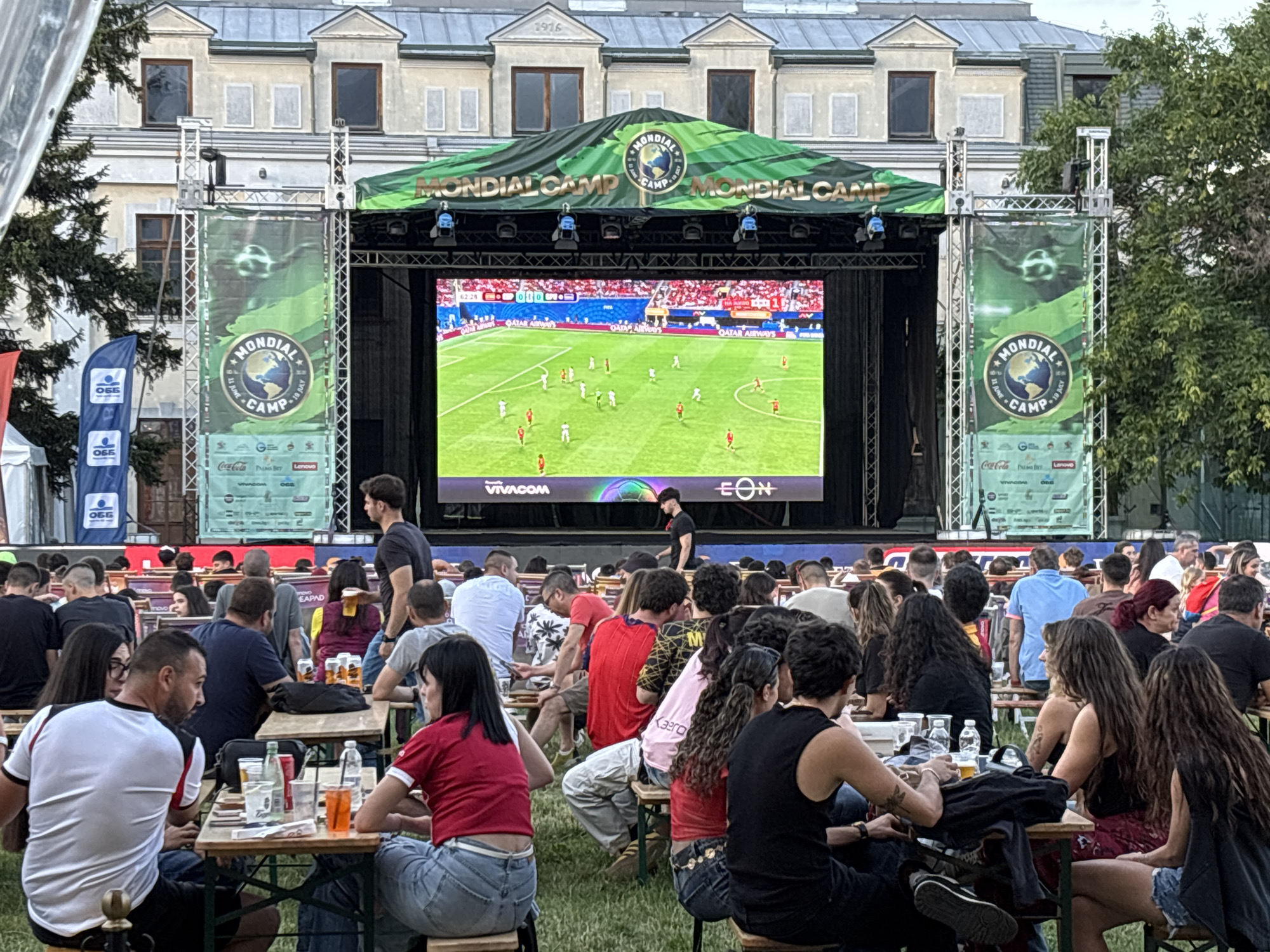 In Sofia, Bulgaria, at the Mondial Camp event, a large outdoor screen displays a soccer match. A crowd of people is gathered, sitting at picnic tables in front of the screen. The scene is set outdoors, with trees visible in the background. The overall atmosphere appears to be casual and festive, with attendees enjoying the game together.