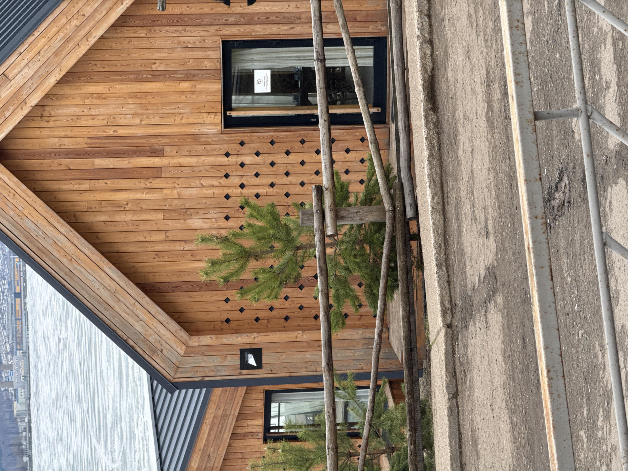 The image presents a view of a wooden building in what is possibly Votkinsk, Russia. The structure is clad in horizontal wooden planks, with a design incorporating black diamond-shaped inlays. It features a window with a black frame and a sign inside. To the side, there is a concrete structure and a metal railing. There is sparse foliage, including small pine trees, obscuring the view of the building. The surrounding environment appears to include a body of water.