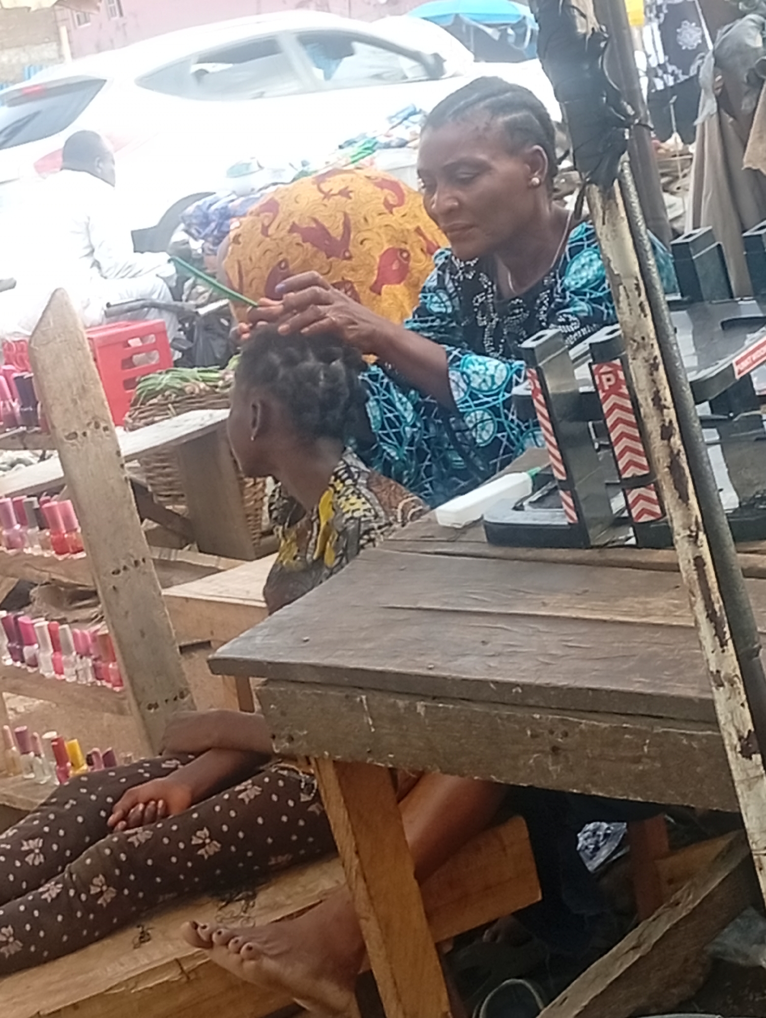In Sabon Tasha, Nigeria, a woman braids another woman's hair in an outdoor market setting. The braider, wearing a blue and black patterned top, is focused on her task. Her client sits on a wooden bench, wearing a yellow patterned top and brown floral pants, with her legs extended. A table cluttered with beauty supplies and a display of nail polish jars are nearby. A white car is visible in the background.