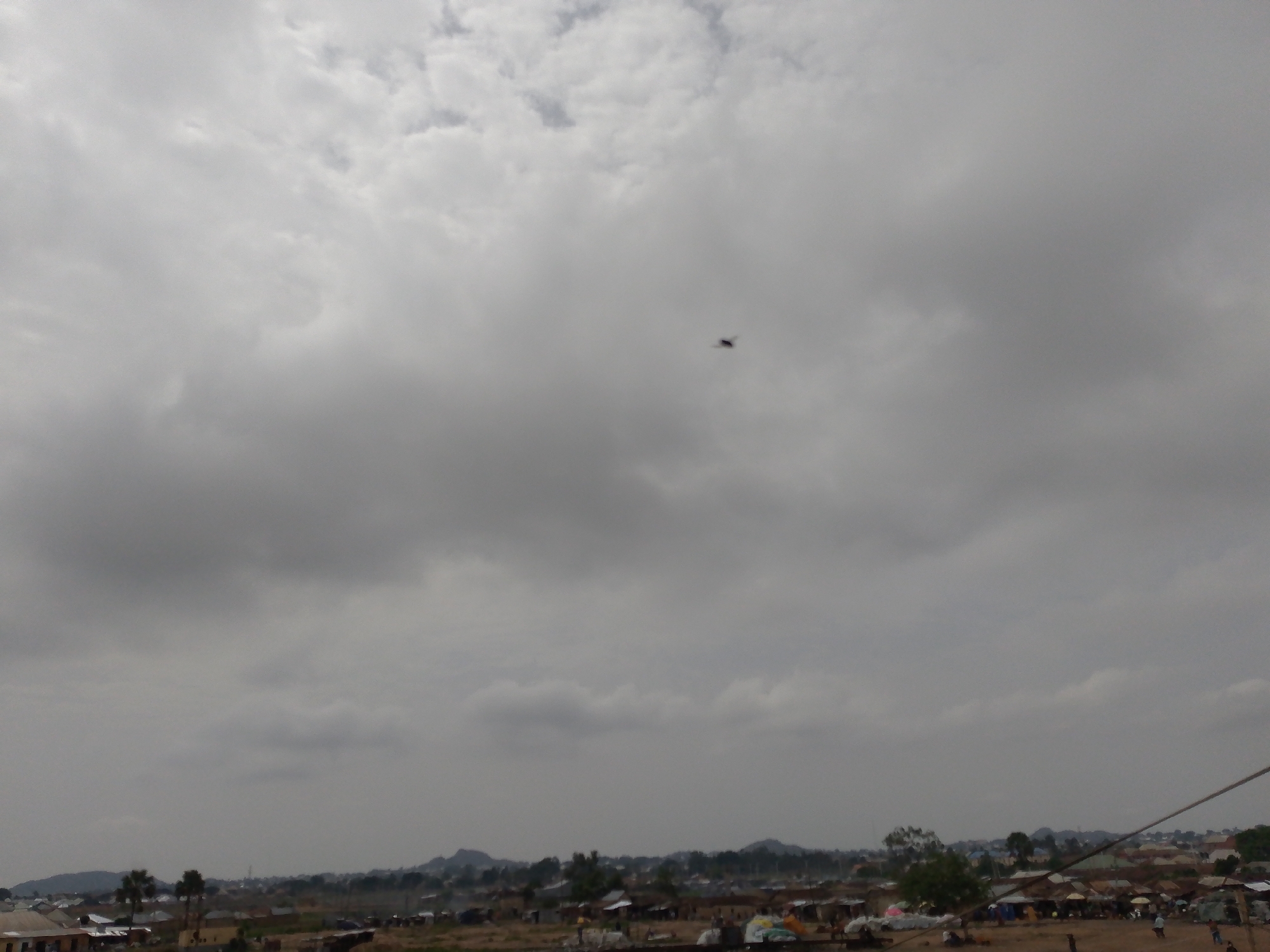 The scene is an overcast day in what appears to be a poorer neighborhood or rural area of Bukuru, Nigeria. Low-lying buildings and dwellings are scattered across the landscape in the foreground, with some trees interspersed. Hills or small mountains are visible in the distance. In the sky, a dark silhouette that appears to be a helicopter flies high above the scene. There are a few figures visible on the ground, interacting or moving around the buildings.