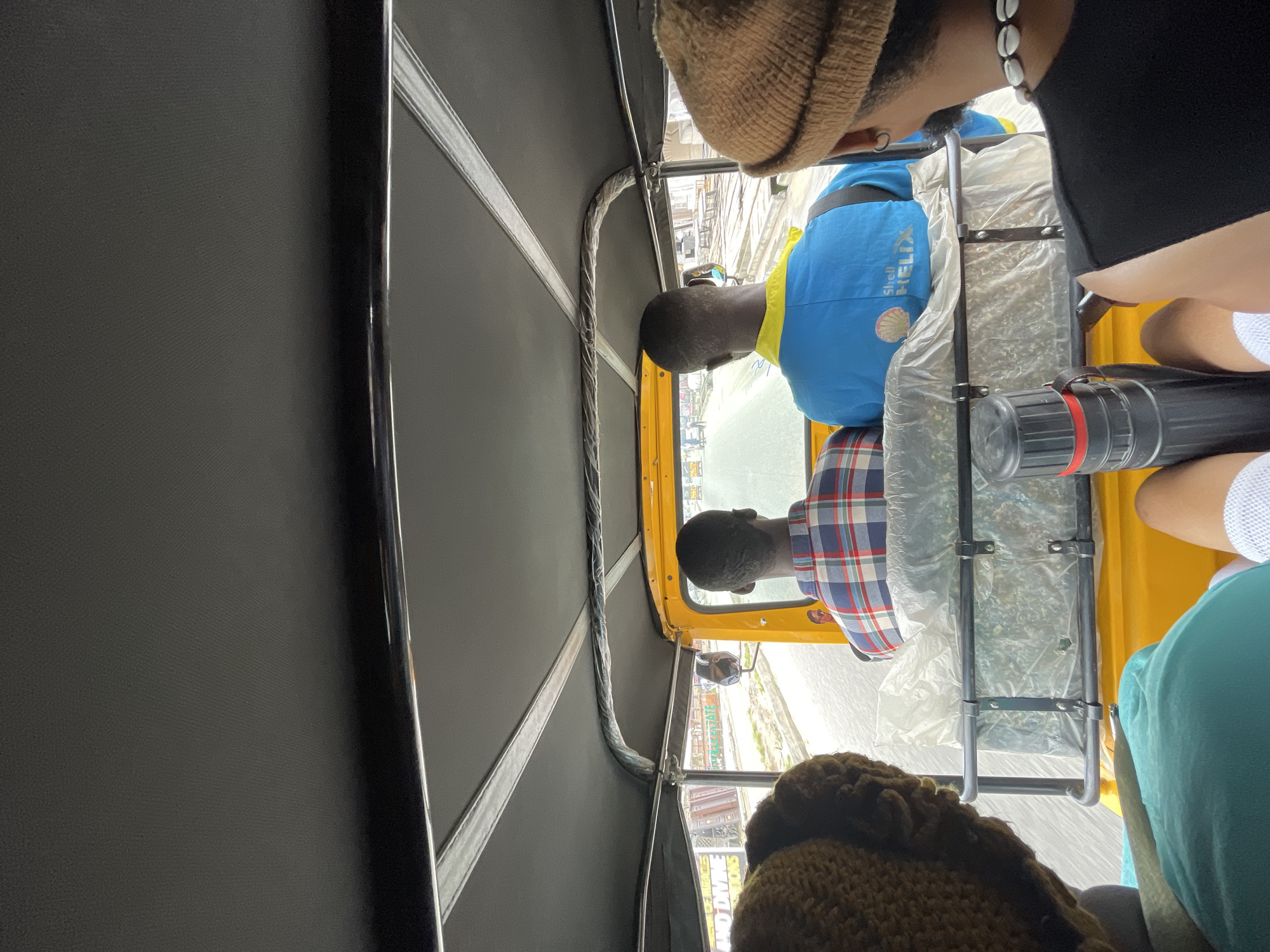 The interior of an auto rickshaw (tuk-tuk) in Ado, Nigeria is shown. Three passengers are visible. One passenger is wearing a brown hat and a necklace with shells, while another is wearing a blue t-shirt with "Shell Helix" printed on it. A third passenger is wearing a turquoise t-shirt. A black canister with a red stripe is sitting on the seat. The rickshaw has a black interior and yellow exterior with a partially visible cityscape outside.