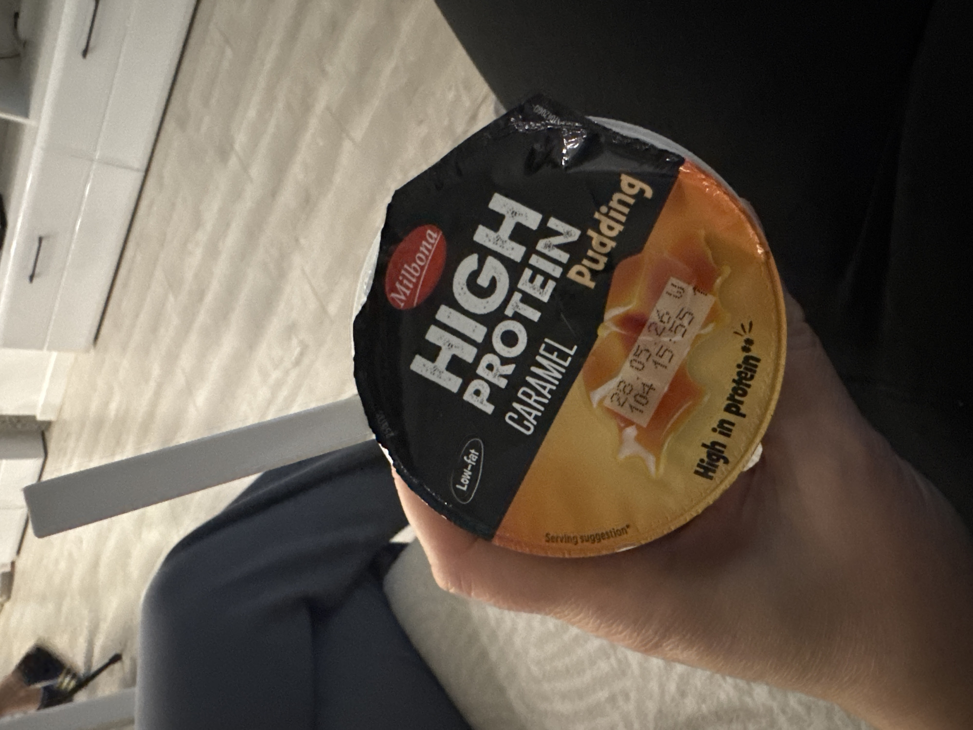 A person holds a Milbona High Protein Caramel Pudding in their hand. The pudding container has a black lid with white text and a caramel-colored base. The expiration date "28.05.26" is visible on the container. The person is seated. The background is a blurry combination of furniture and flooring. The location is unknown and not specified in the image.