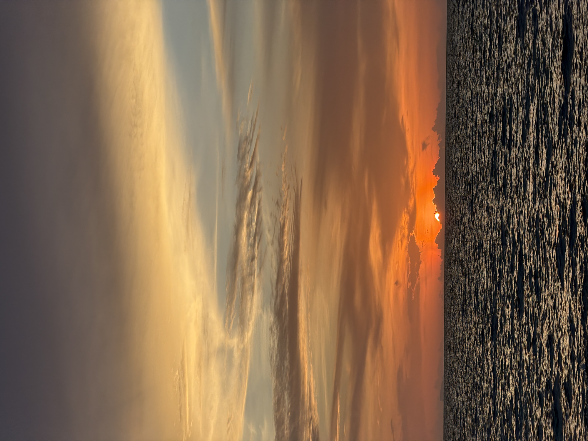 The image depicts a sunset over the ocean. The sky is filled with a gradient of colors, ranging from deep purple at the top to vibrant oranges and yellows closer to the horizon. Wispy clouds are scattered across the sky, catching the warm light of the setting sun. The sun itself is partially obscured by clouds, creating a dramatic glow. Below the sky is the dark, rippling surface of the ocean. The water reflects the colors of the sky, with patches of lighter hues interspersed with darker areas. The horizon line is clearly defined, separating the sky and the sea. The overall impression is one of a serene and beautiful natural landscape. The location is identified as the Ko Samui District in Thailand, suggesting a tropical coastal setting. There are no people or man-made structures visible in the image.